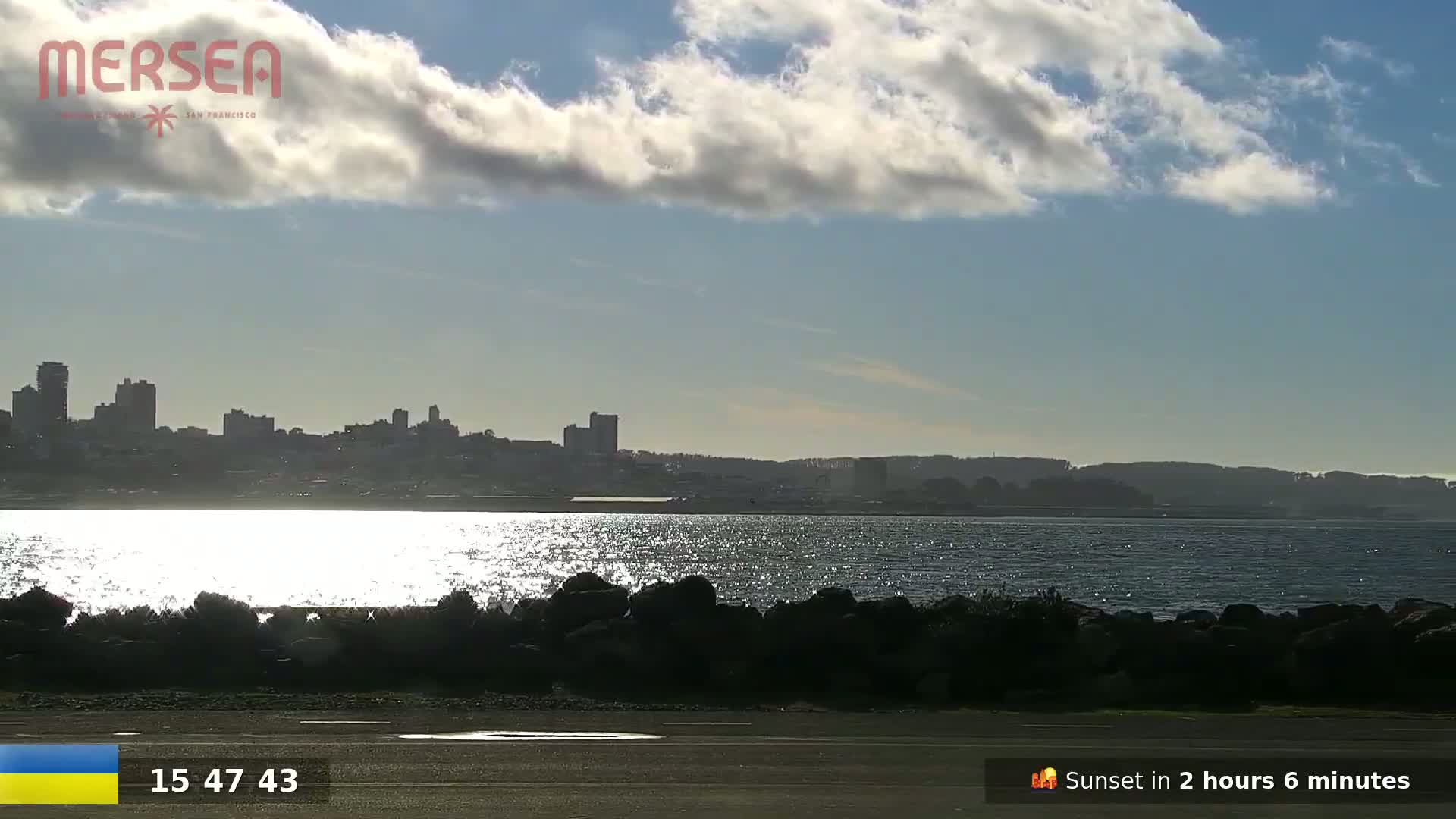 A city skyline is visible across a body of sparkling water under a partly cloudy sky.