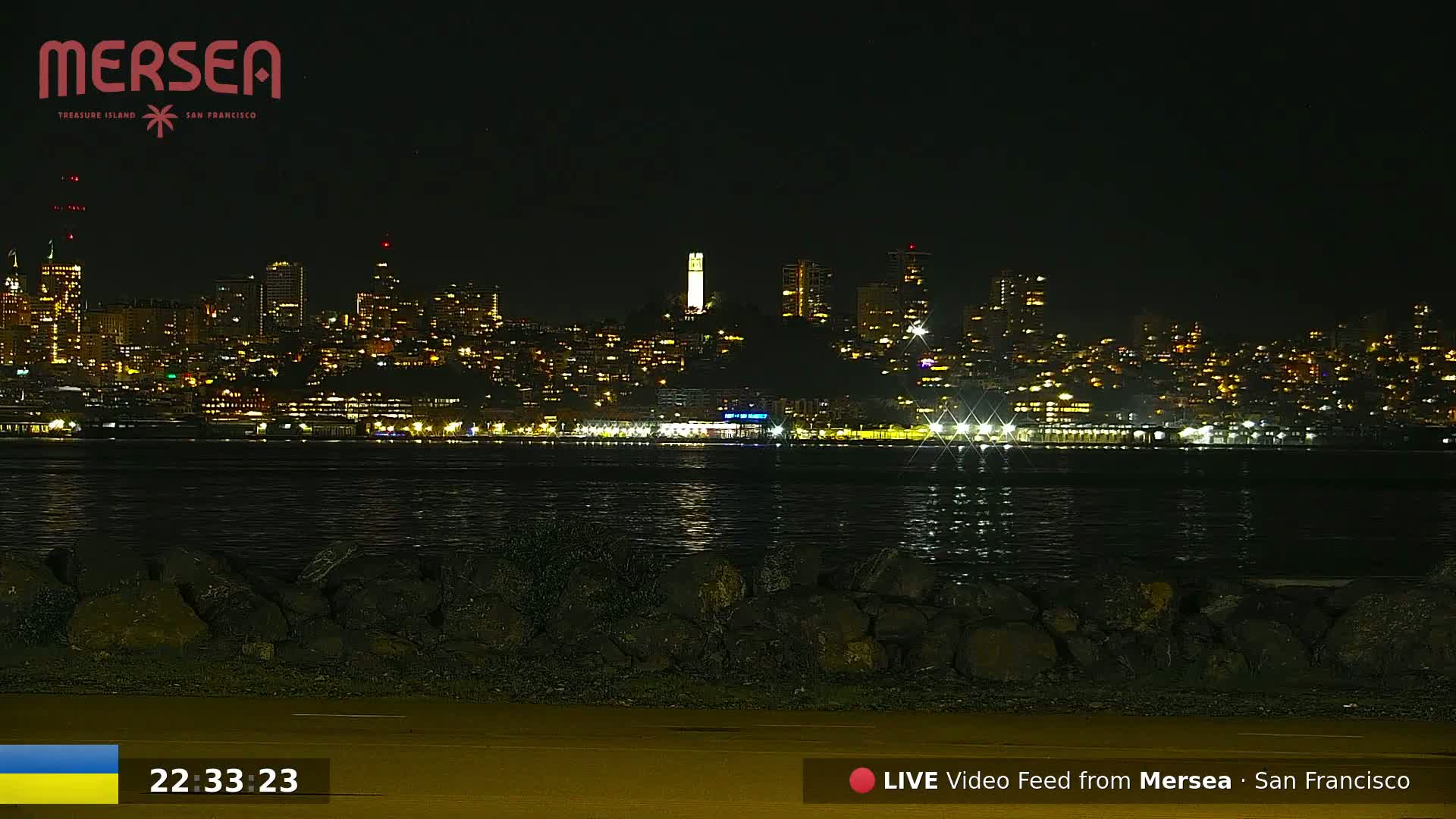 A nighttime view of a city skyline across a body of water, with a rocky shoreline in the foreground.
