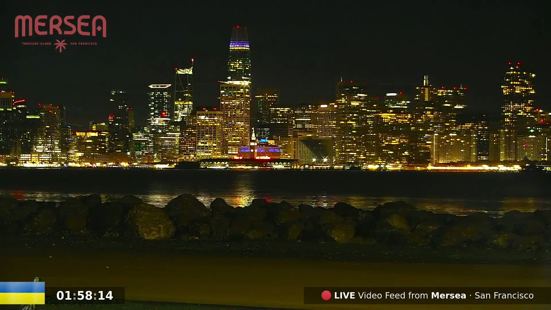 A nighttime view of a brightly lit city skyline across a calm body of water, with dark rocks in the foreground.