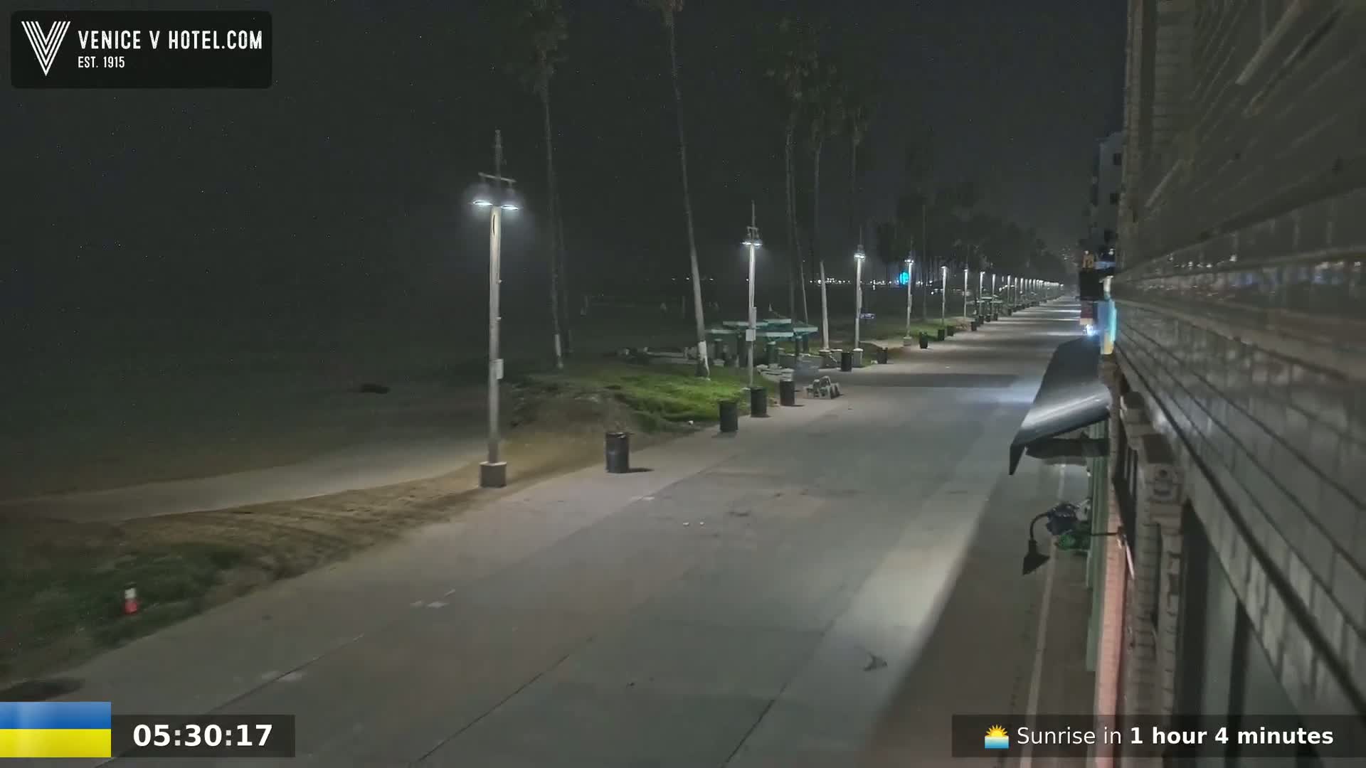 A nighttime view of a mostly empty boardwalk next to a beach, lined with palm trees and streetlights.