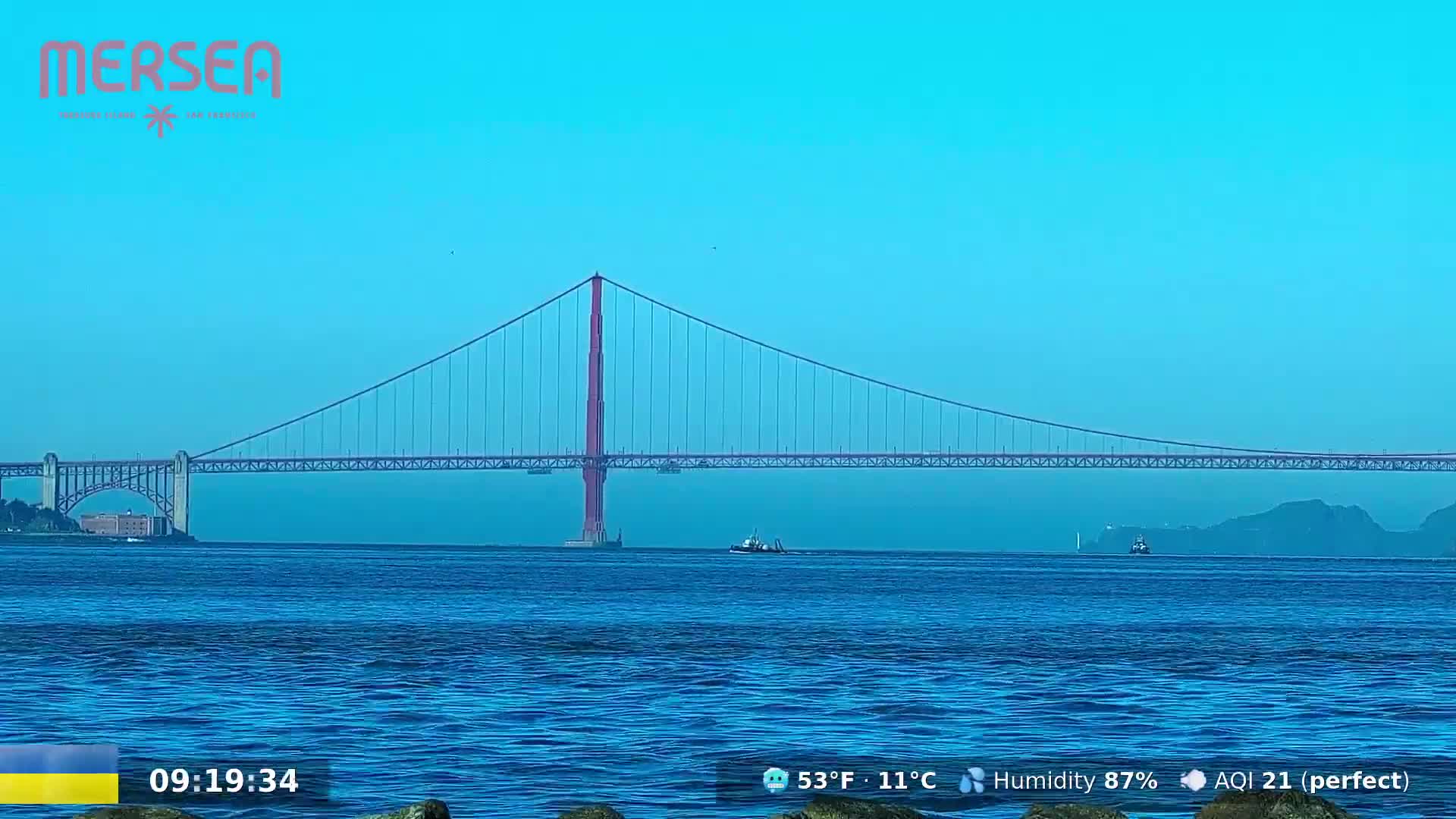 A suspension bridge spans a calm, blue body of water under a clear, light-blue sky.