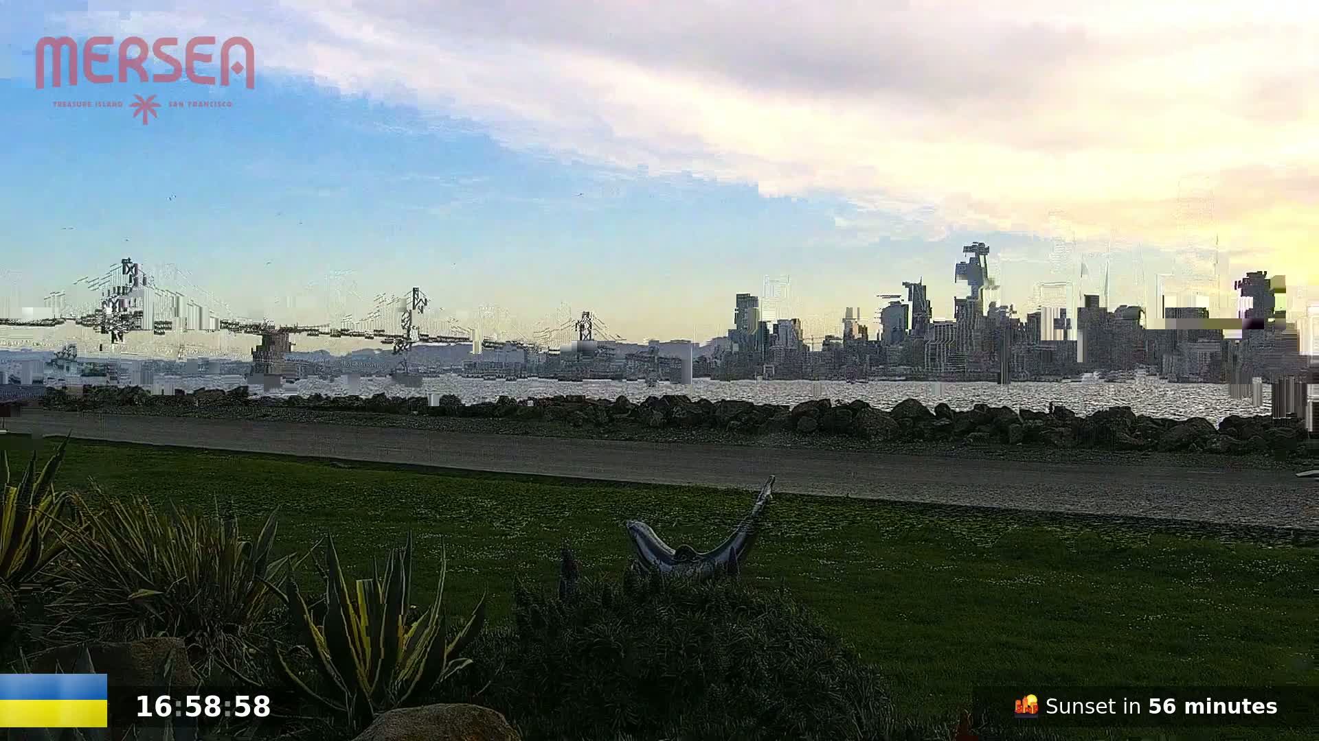 A grassy park overlooks a calm body of water, with a cityscape and bridge visible in the distance under a partly cloudy sky.