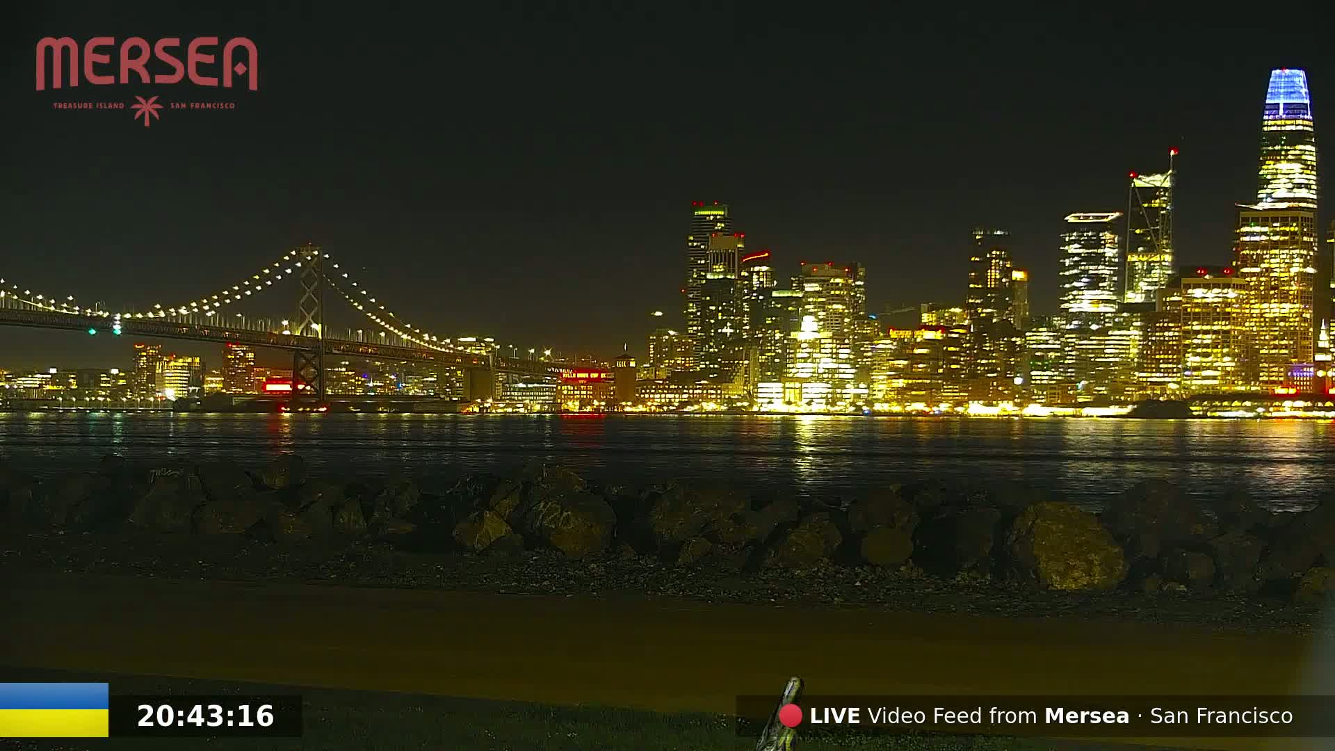 A nighttime view of a brightly lit city skyline across a calm body of water, with a suspension bridge in the foreground.