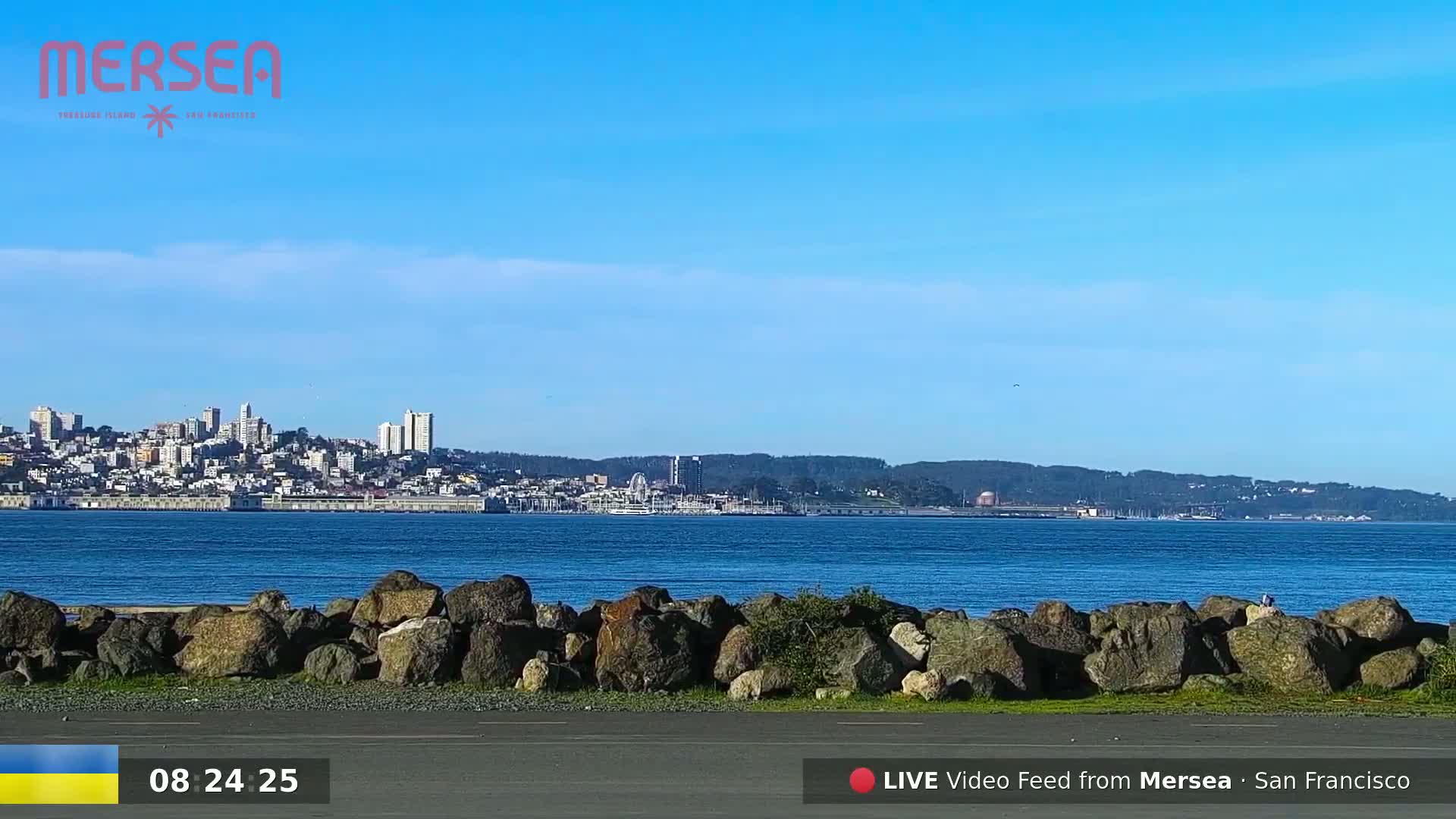 A clear, sunny day reveals a cityscape across a calm body of water, partially obscured by a rocky shoreline in the foreground.