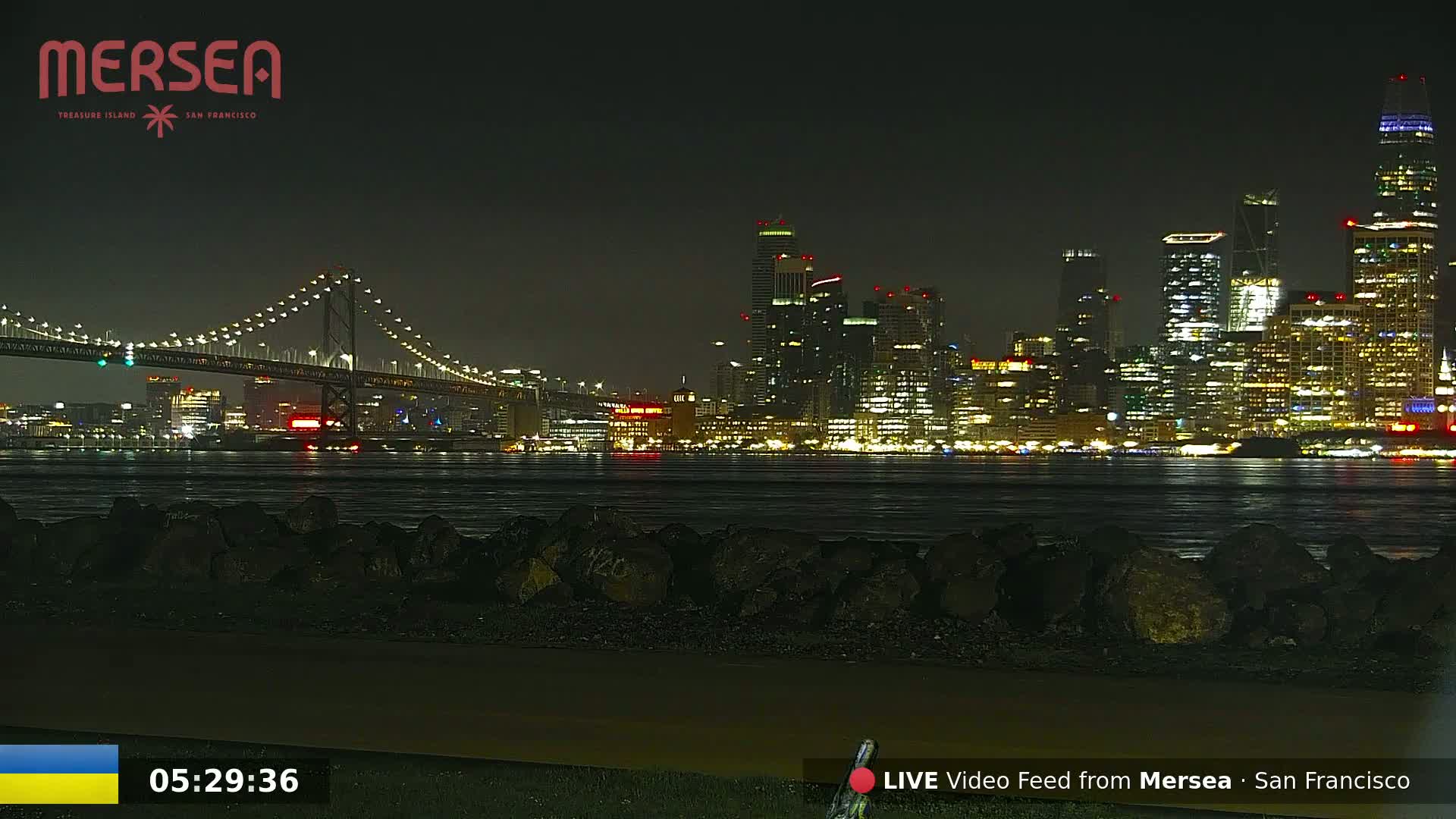 A nighttime view of a city skyline across a body of water, with a bridge illuminated in the foreground under a clear, dark sky.