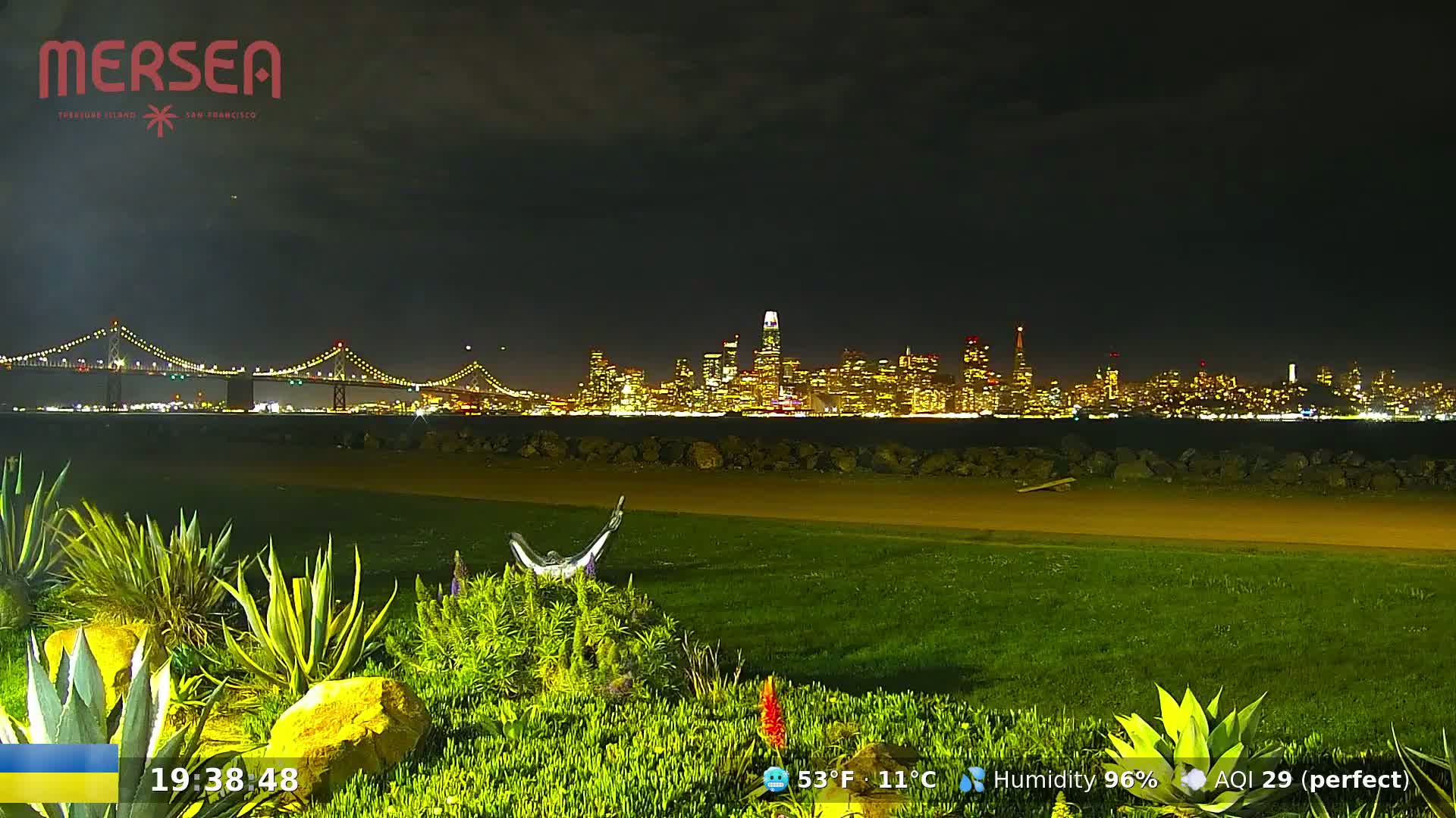 A nighttime view of a city skyline across a body of water, illuminated by city lights, with a bridge in the foreground and lush green landscaping in the immediate foreground.