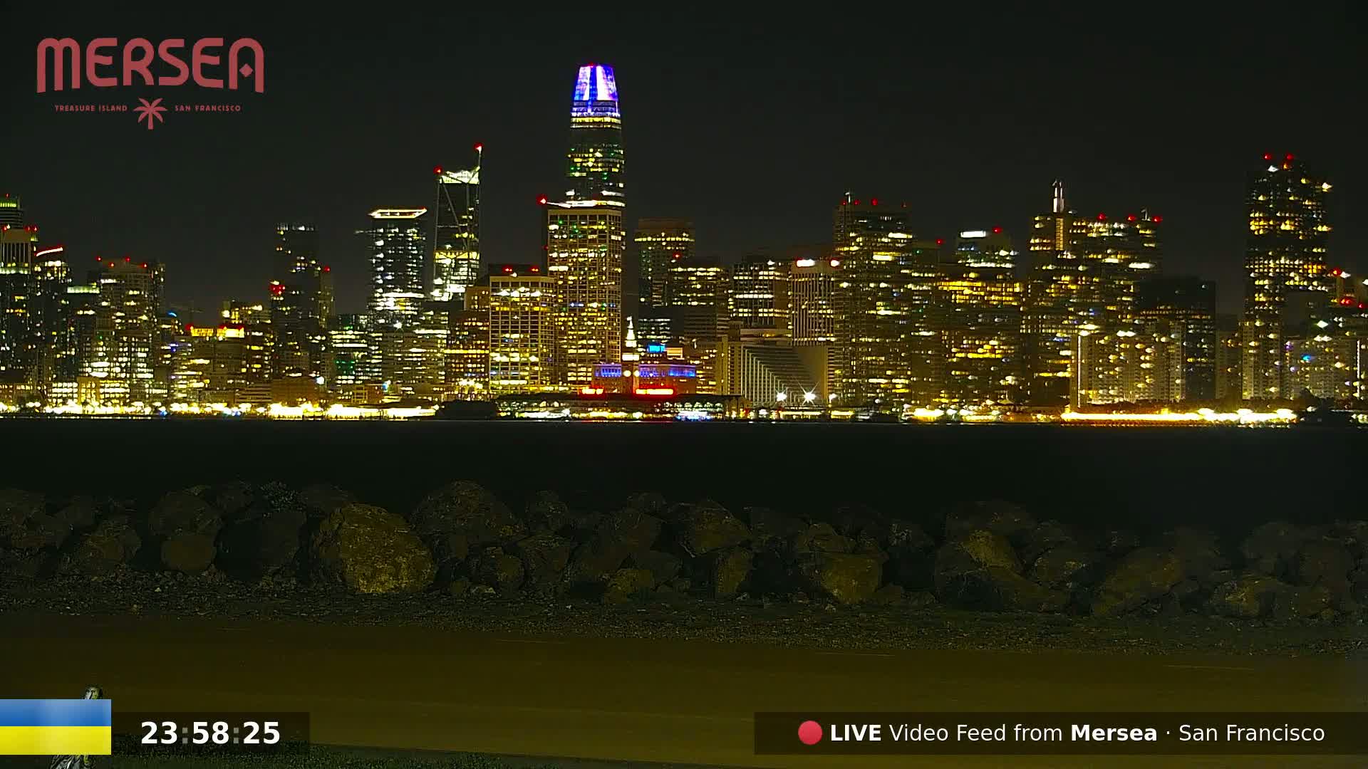 A brightly lit San Francisco city skyline at night is viewed from across a body of water, with a rocky shoreline in the foreground.