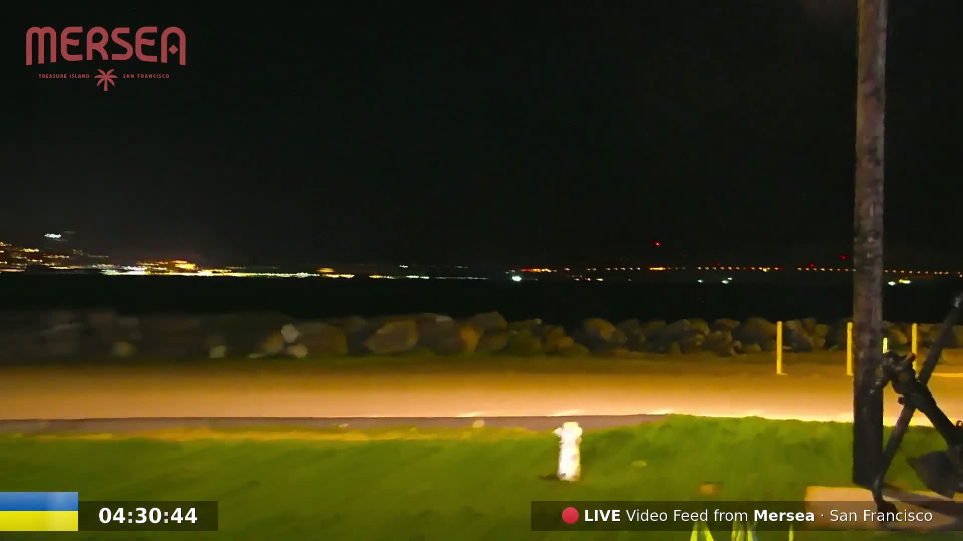 A nighttime view of a city skyline across a body of water, seen from a grassy area with a paved path and rock barrier in the foreground.