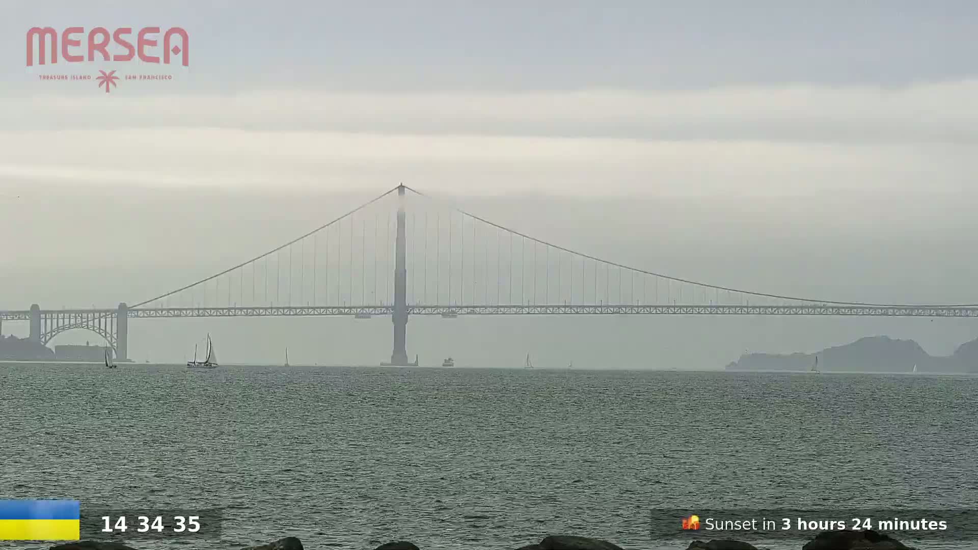 A large suspension bridge spans a hazy, gray body of water with several sailboats visible in the distance.