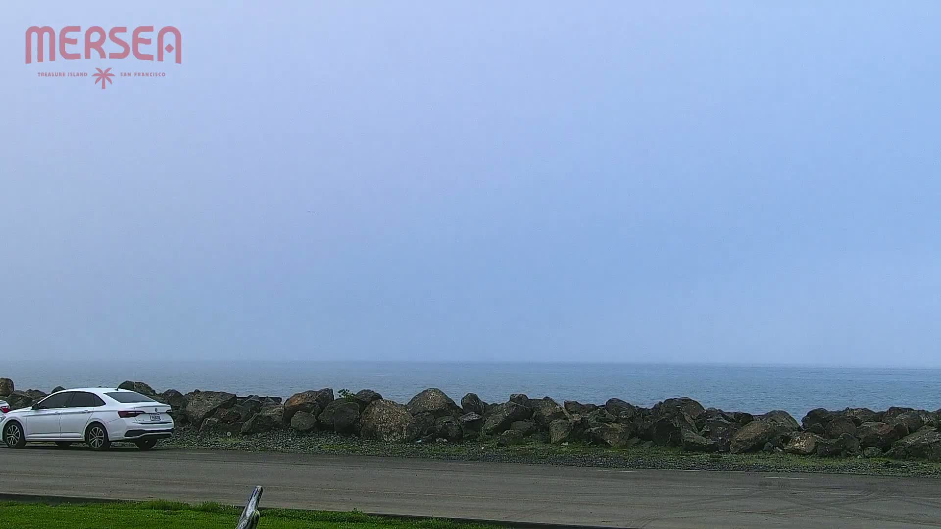A white car is parked near a rocky shoreline overlooking a calm, hazy ocean.