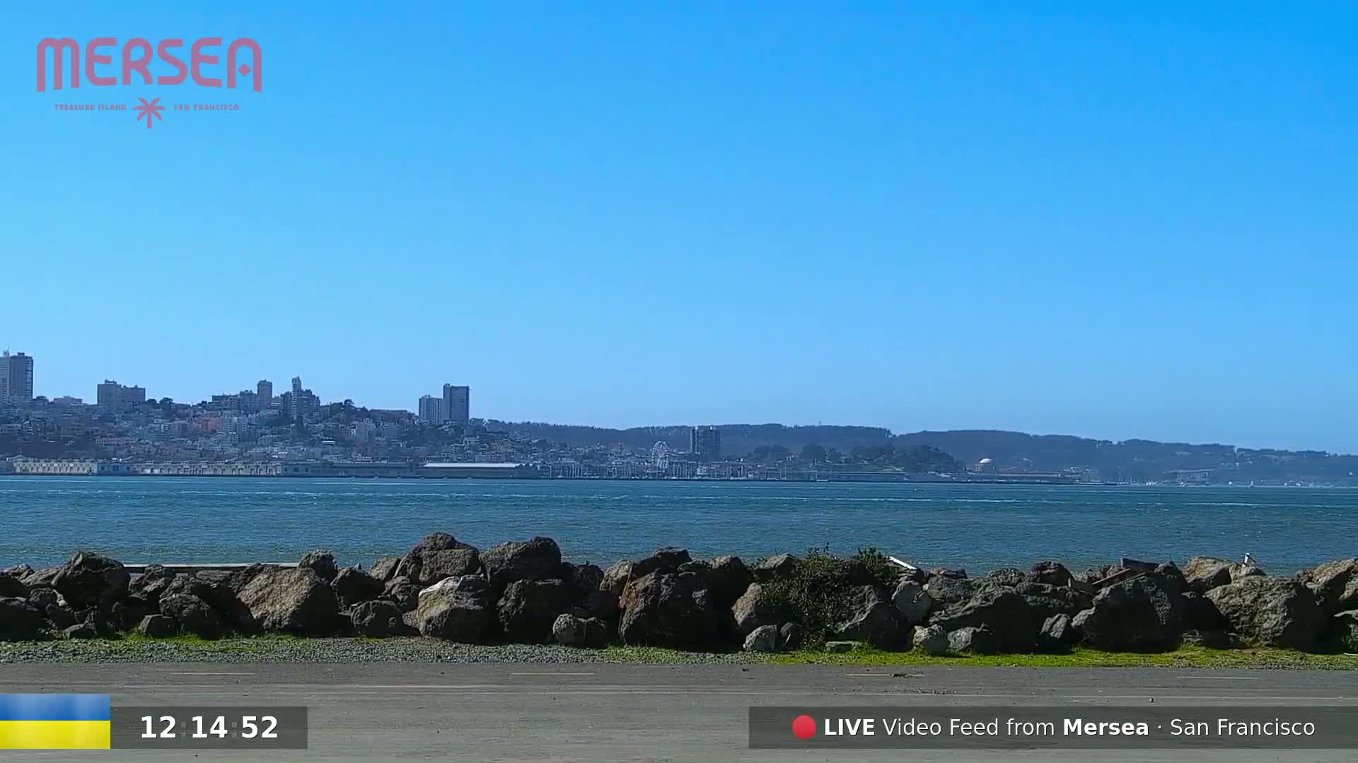 A clear, sunny day shows a cityscape across a body of water, with large rocks in the foreground.