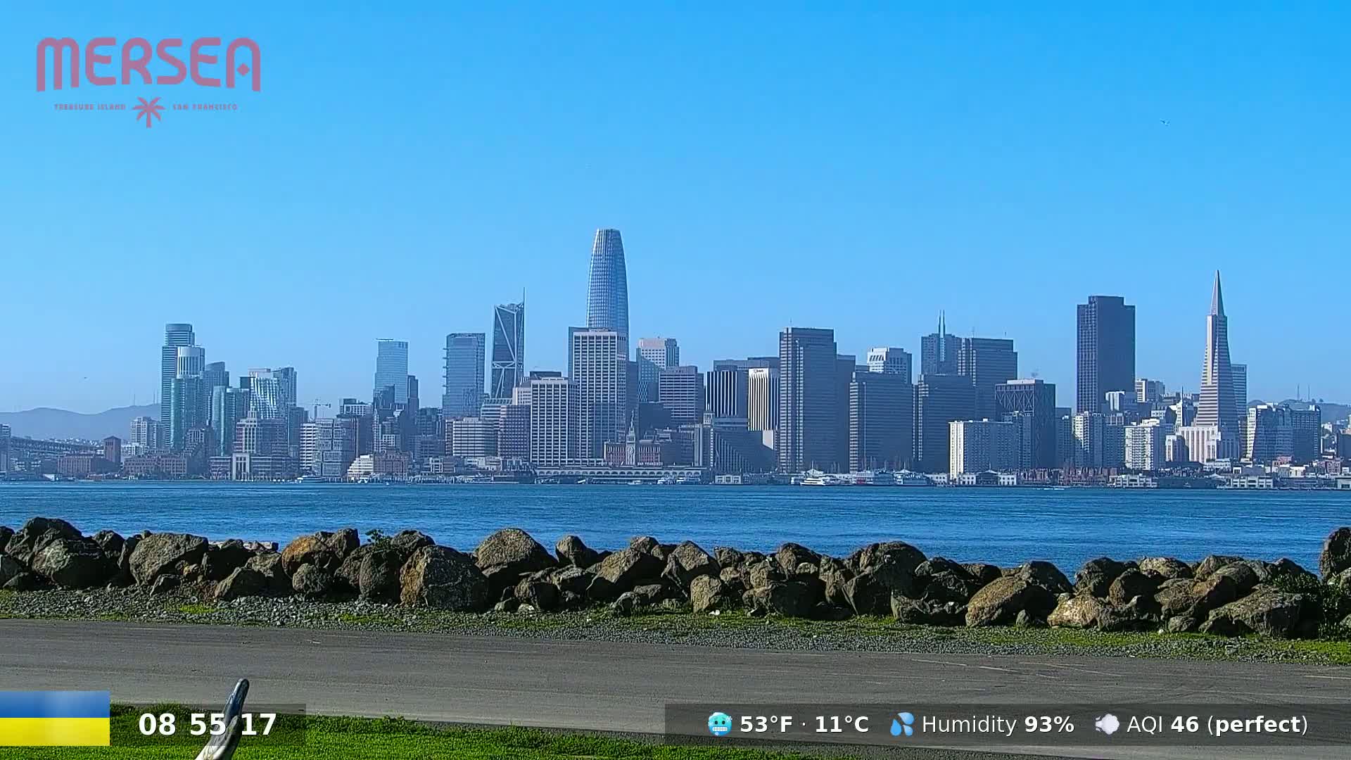 A city skyline is visible across a body of water, on a clear, sunny day with high humidity.