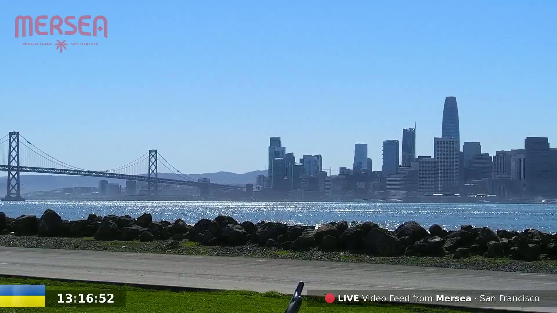 A sunny day shows a suspension bridge, a rocky shoreline, and the San Francisco skyline across a sparkling body of water.