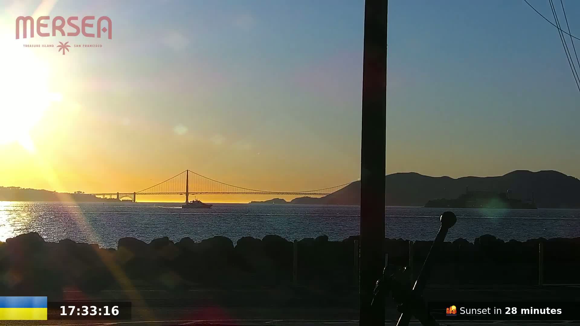 A sunset illuminates a suspension bridge and a body of water with a boat and Alcatraz Island in the distance.