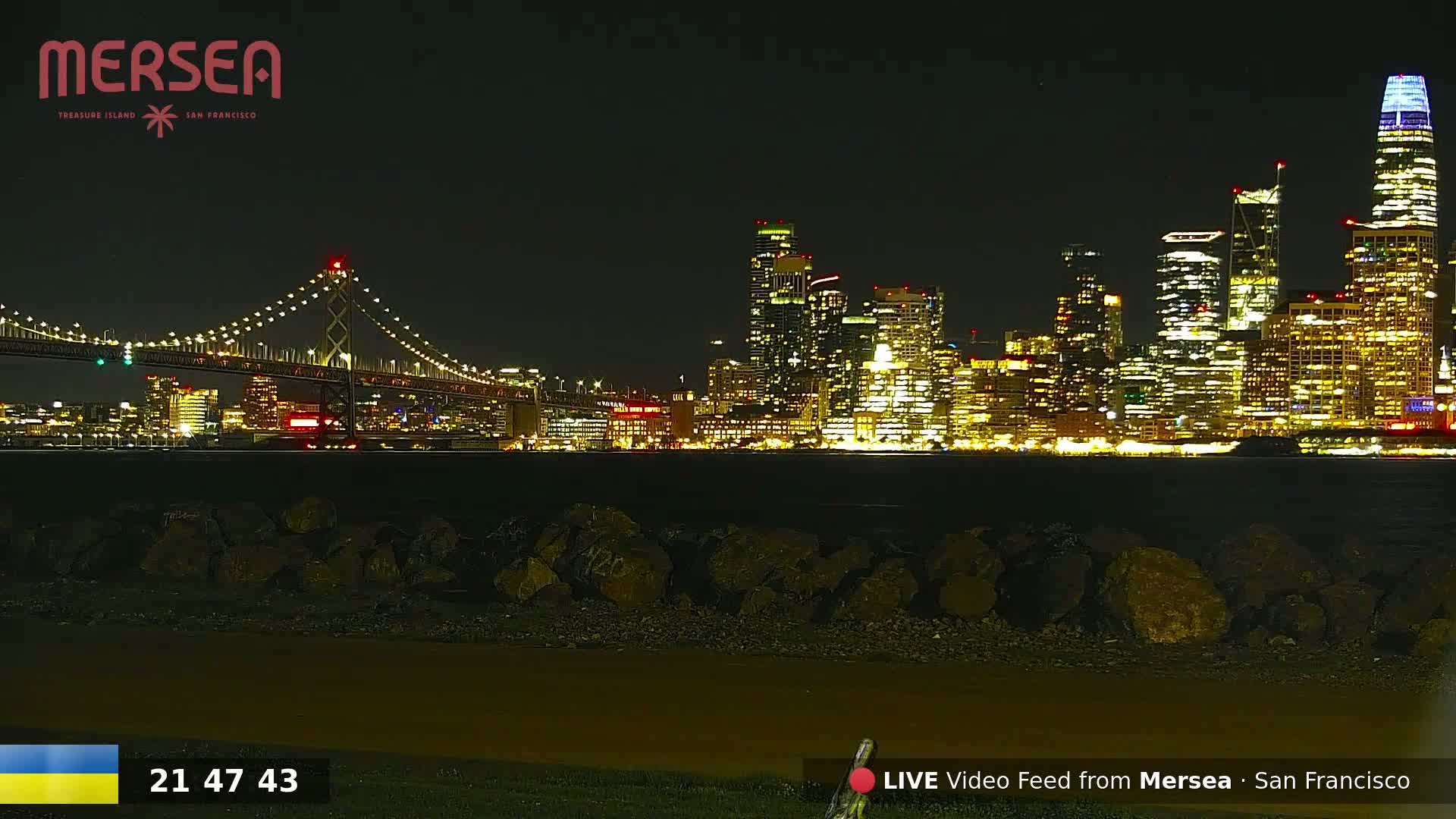 A nighttime view of a brightly lit city skyline across a body of water, with a suspension bridge in the foreground.