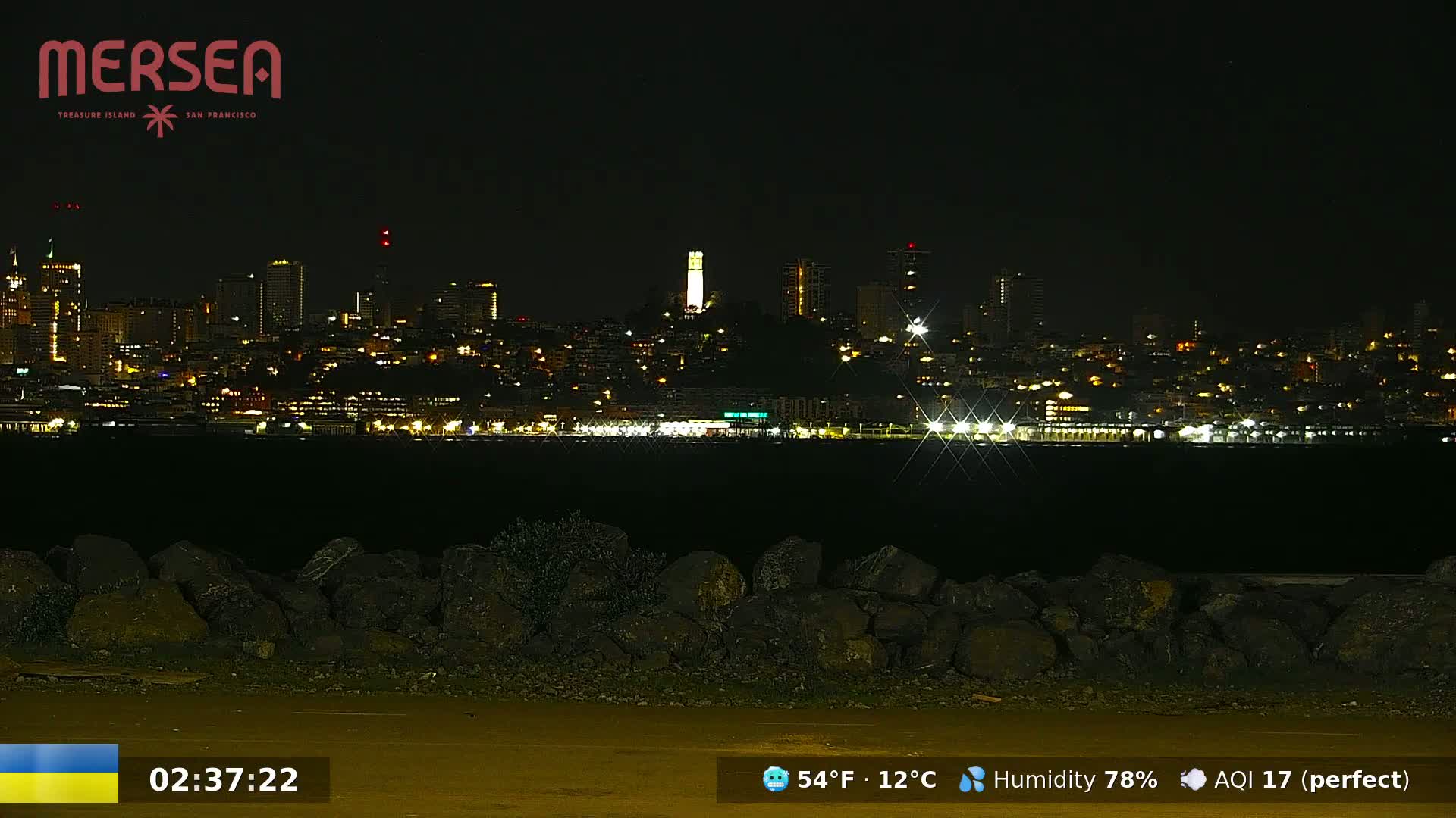 A nighttime view of a city skyline across a body of water, with a rocky shore in the foreground and a clear sky.