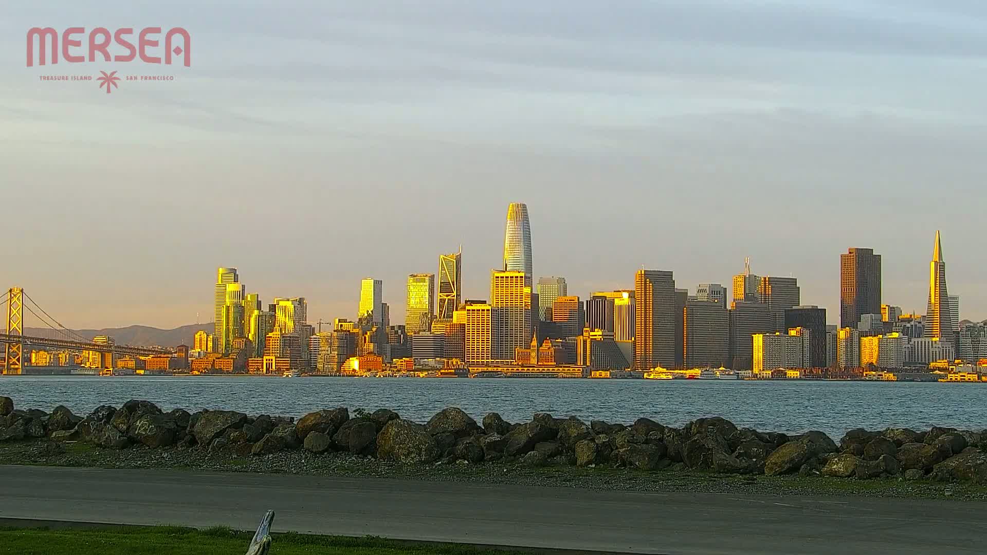 A city skyline, bathed in the golden light of sunset, is viewed from across a body of water, with a rock wall in the foreground.
