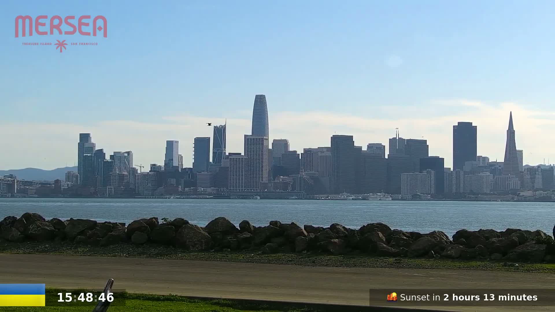 A cityscape is visible across a body of water on a clear, sunny day, with a rocky shoreline in the foreground.