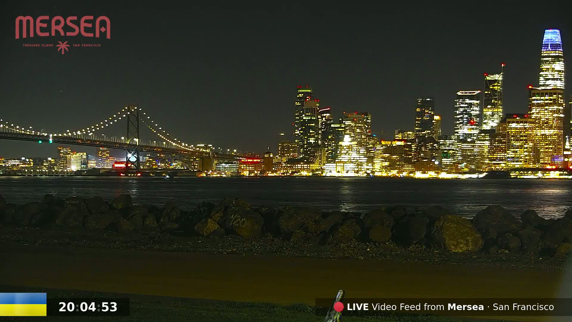 A nighttime view of a brightly lit city skyline across a body of water, with a suspension bridge in the foreground.