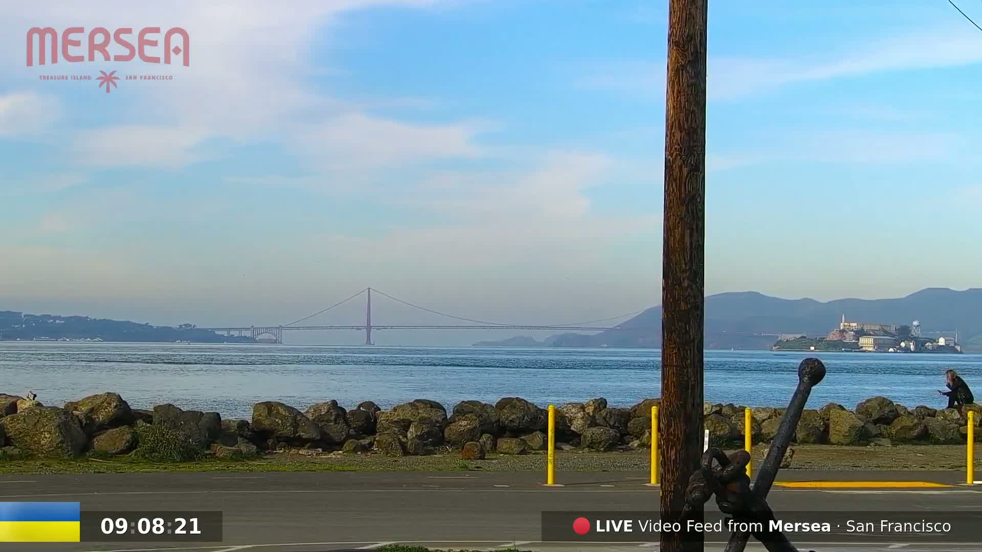 A rocky shoreline overlooks a calm body of water with a suspension bridge and Alcatraz Island in the distance under a clear, bright sky.