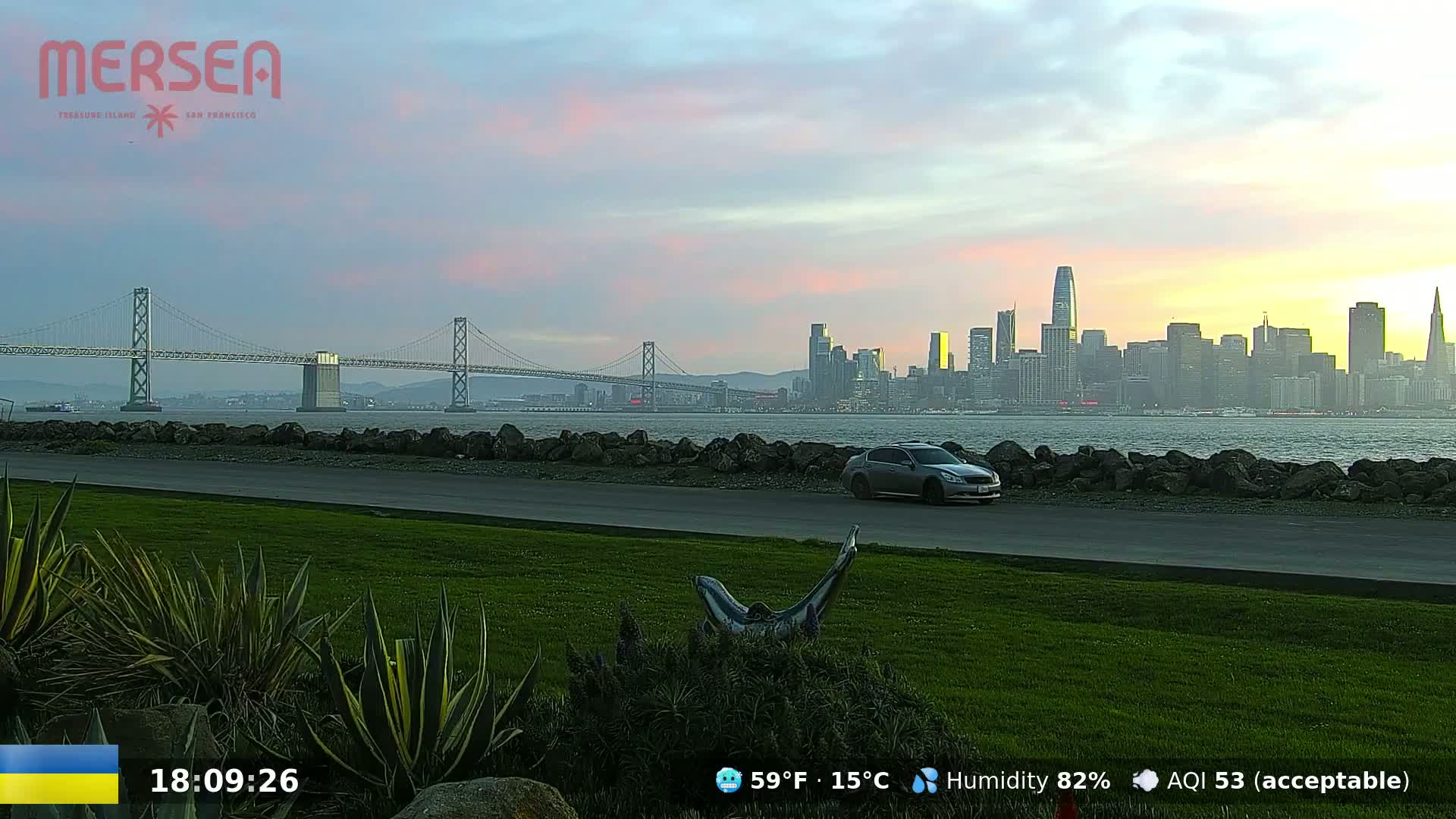A gray car is parked on a grassy area overlooking a body of water, a bridge, and a city skyline at sunset under a partly cloudy sky.