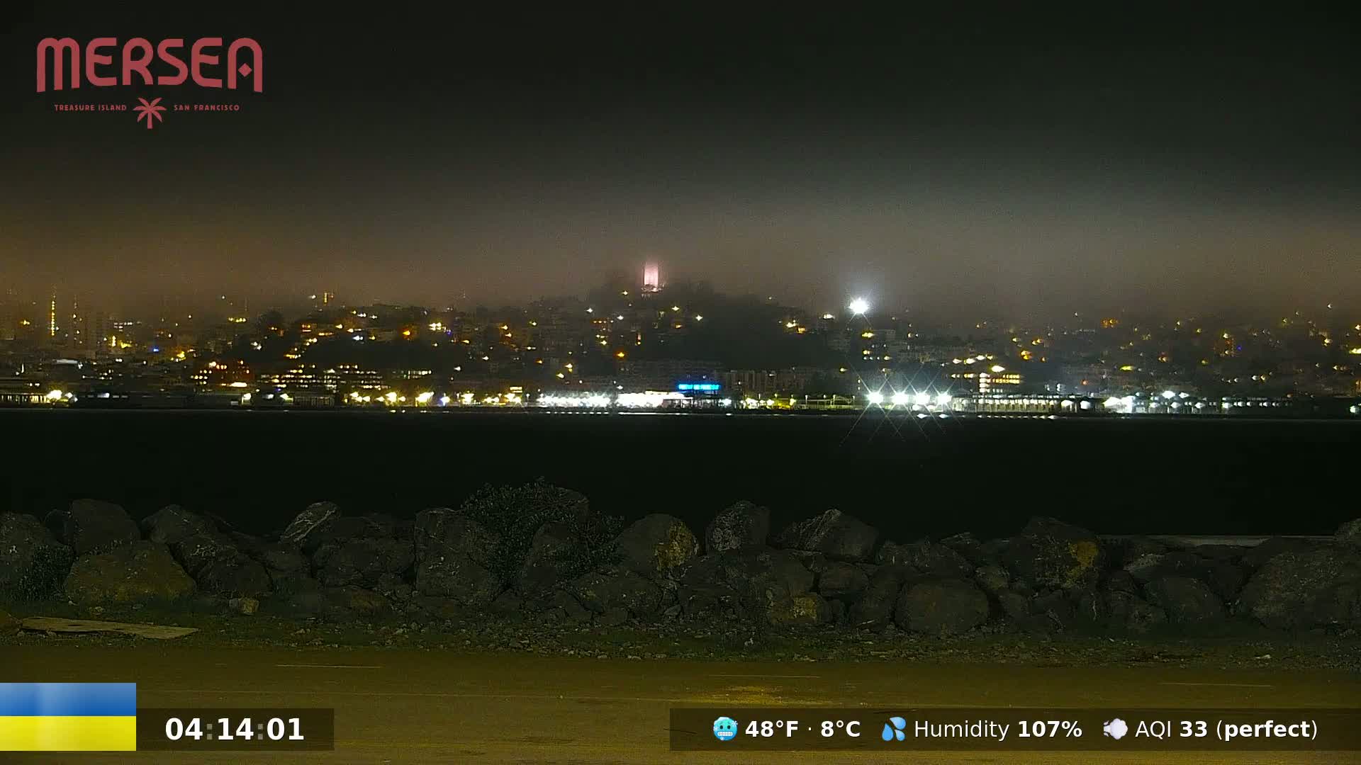A foggy night scene shows a city skyline illuminated by lights across a dark body of water, with a rocky shoreline in the foreground.