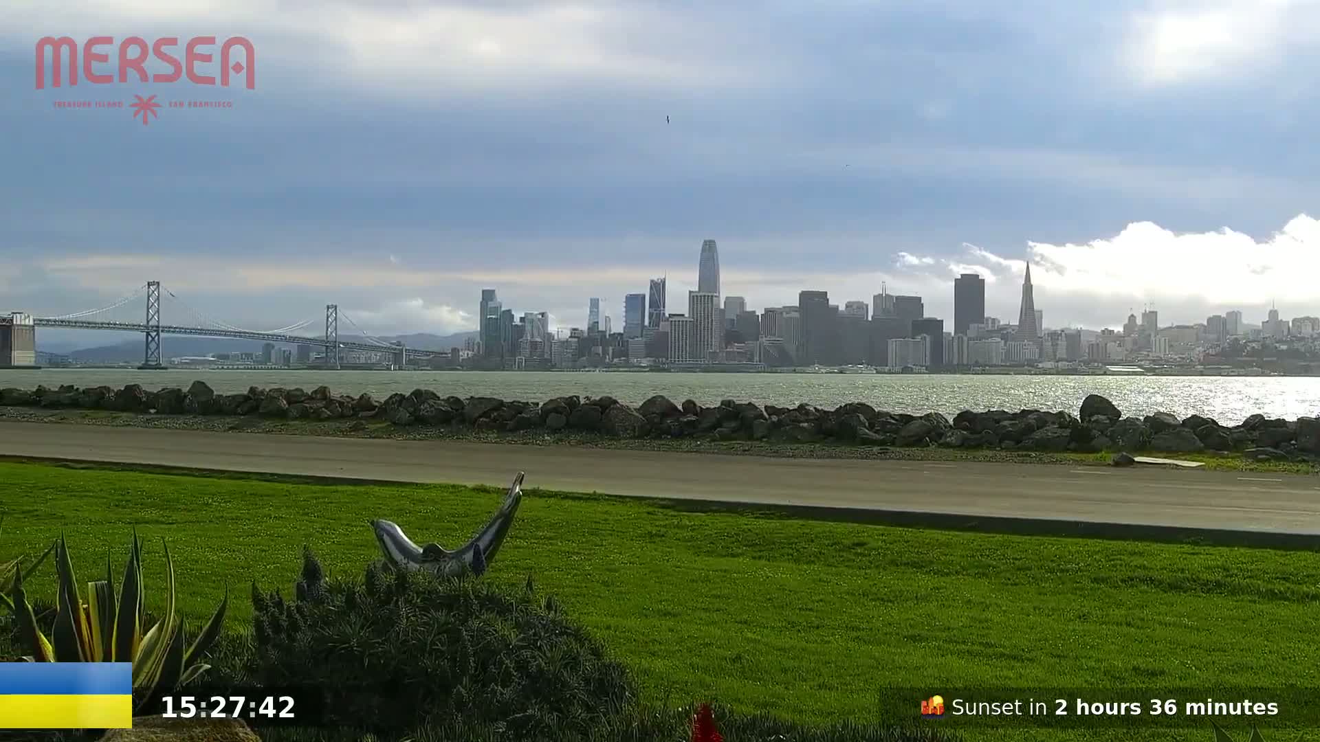 A grassy area with a dolphin statue overlooks a body of water, a bridge, and the San Francisco skyline under a partly cloudy sky.