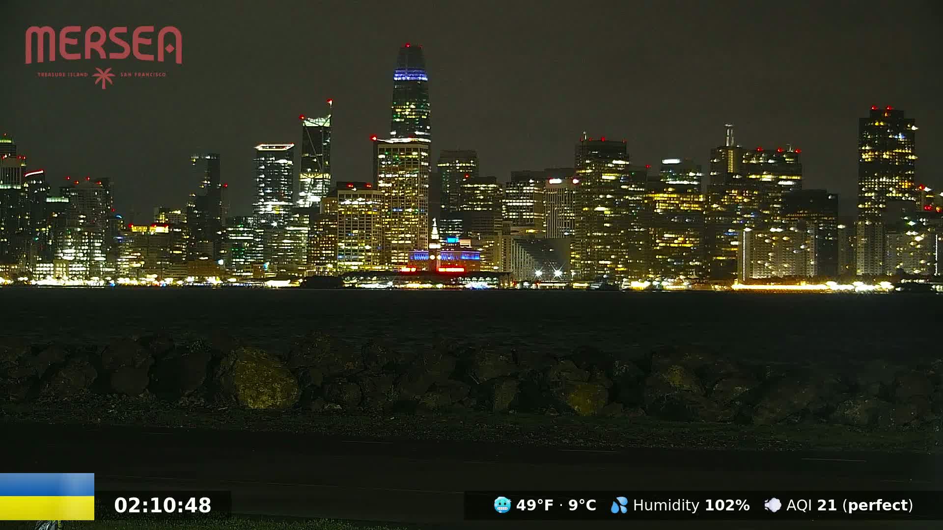A nighttime view of a brightly lit city skyline across a dark body of water, under conditions of 49°F (9°C) and 102% humidity.
