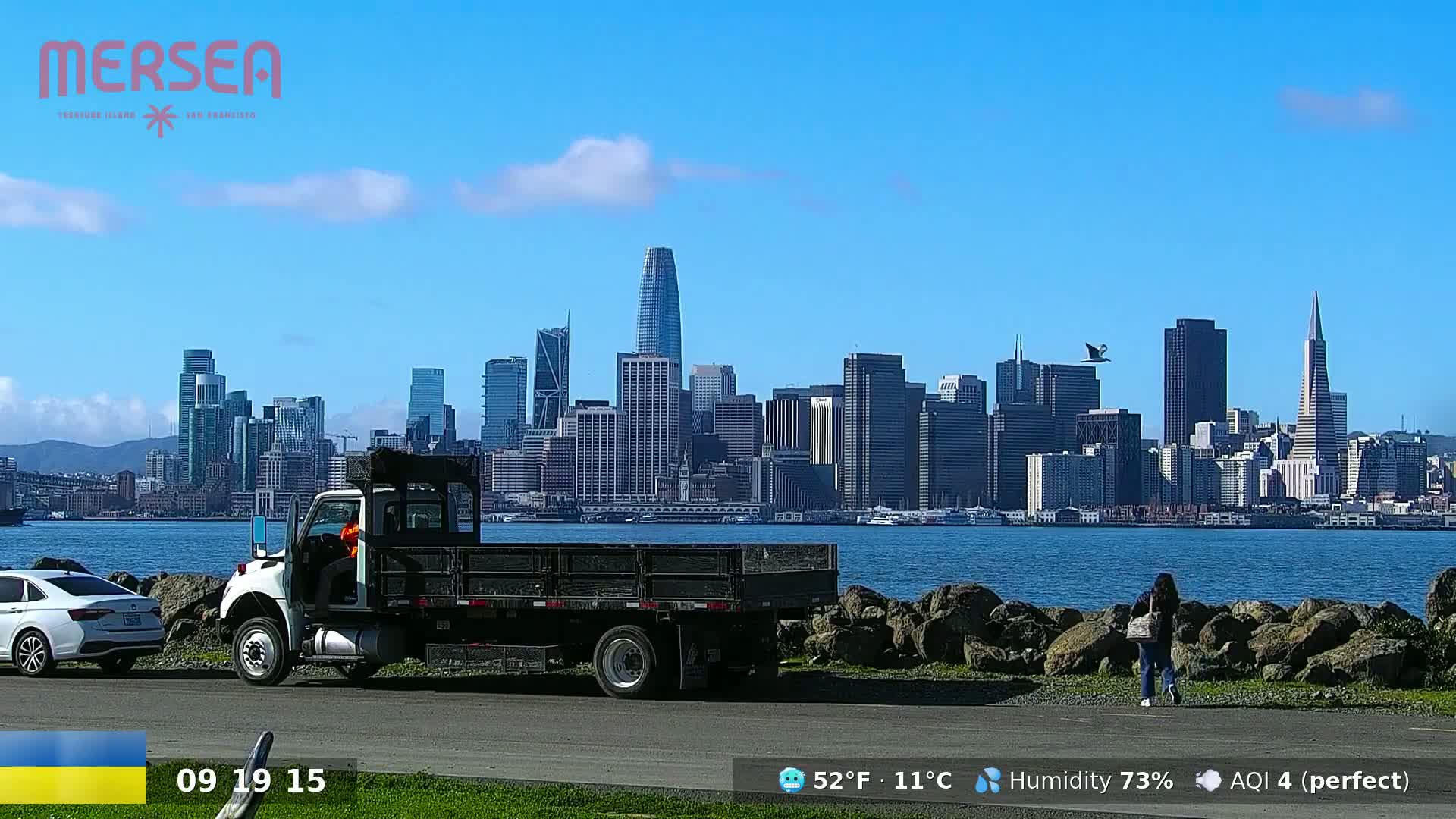 A city skyline is visible across a body of water under a clear blue sky with a few clouds, a truck and a person are seen in the foreground.