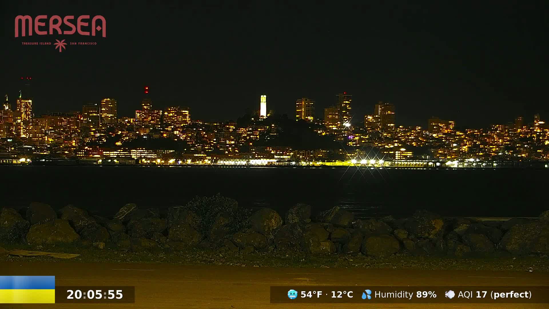 A nighttime view of a brightly lit city skyline across a dark body of water, with a rocky shore in the foreground.