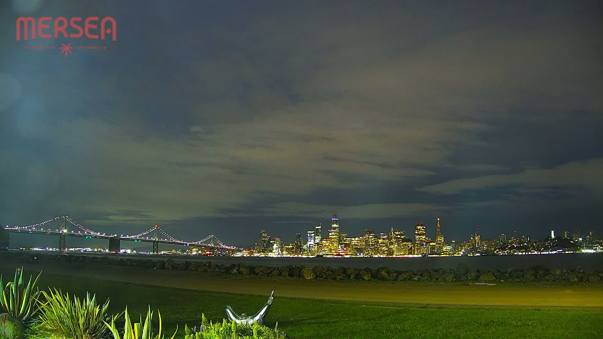 A nighttime view of a city skyline across a body of water, under a cloudy sky, with a bridge in the foreground.