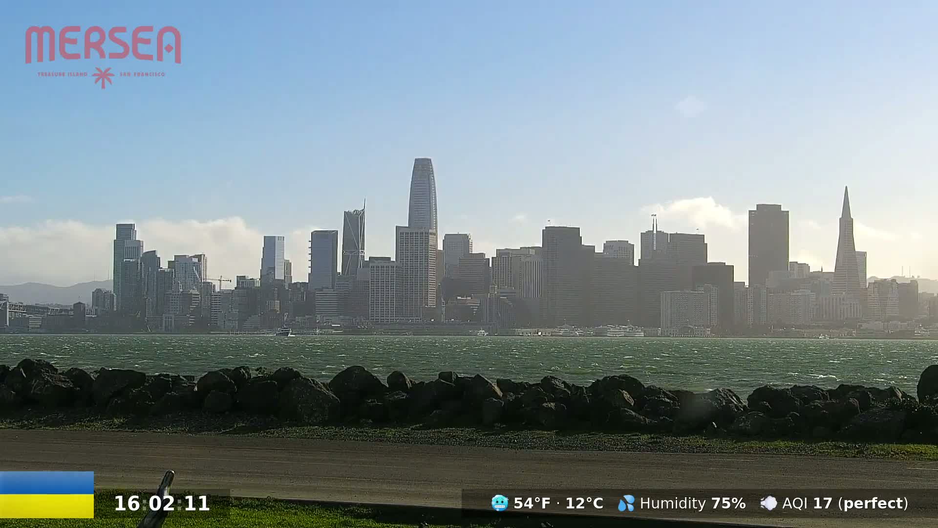 A city skyline with numerous skyscrapers is visible across a body of water with a slightly choppy surface under a mostly clear, sunny sky.