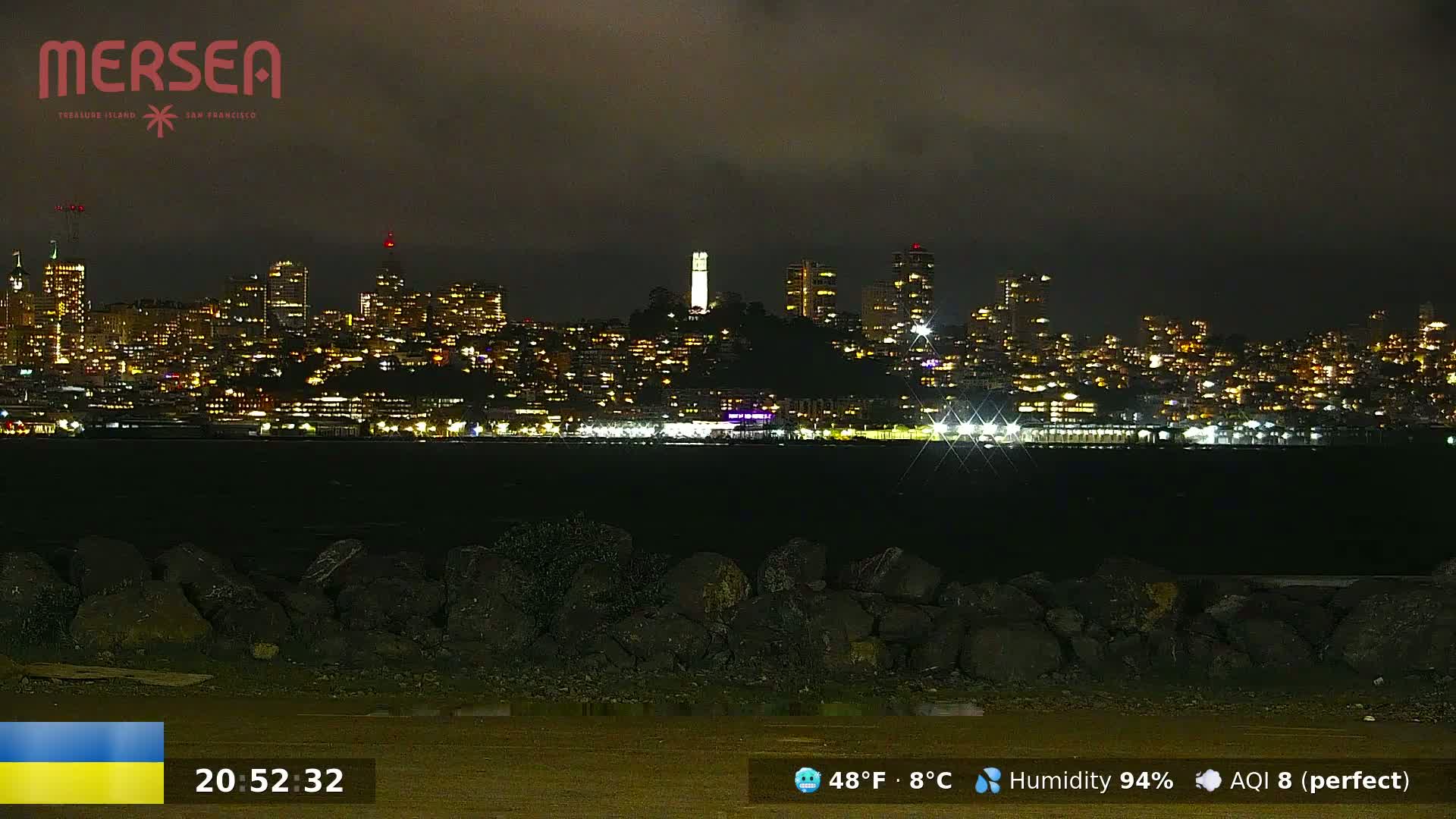A nighttime view of a city skyline illuminated by lights across a dark body of water, under a cloudy sky with a temperature of 48°F (8°C) and 94% humidity.