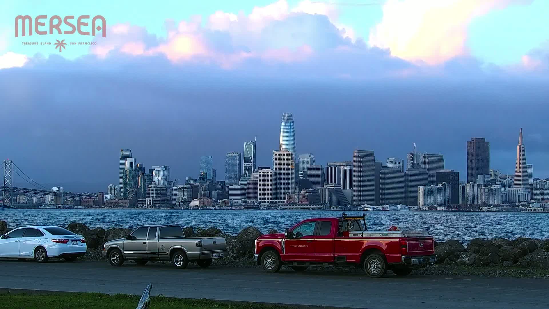 A city skyline is seen across a body of water from a vantage point where two pickup trucks and a sedan are parked, under a cloudy, dusky sky.