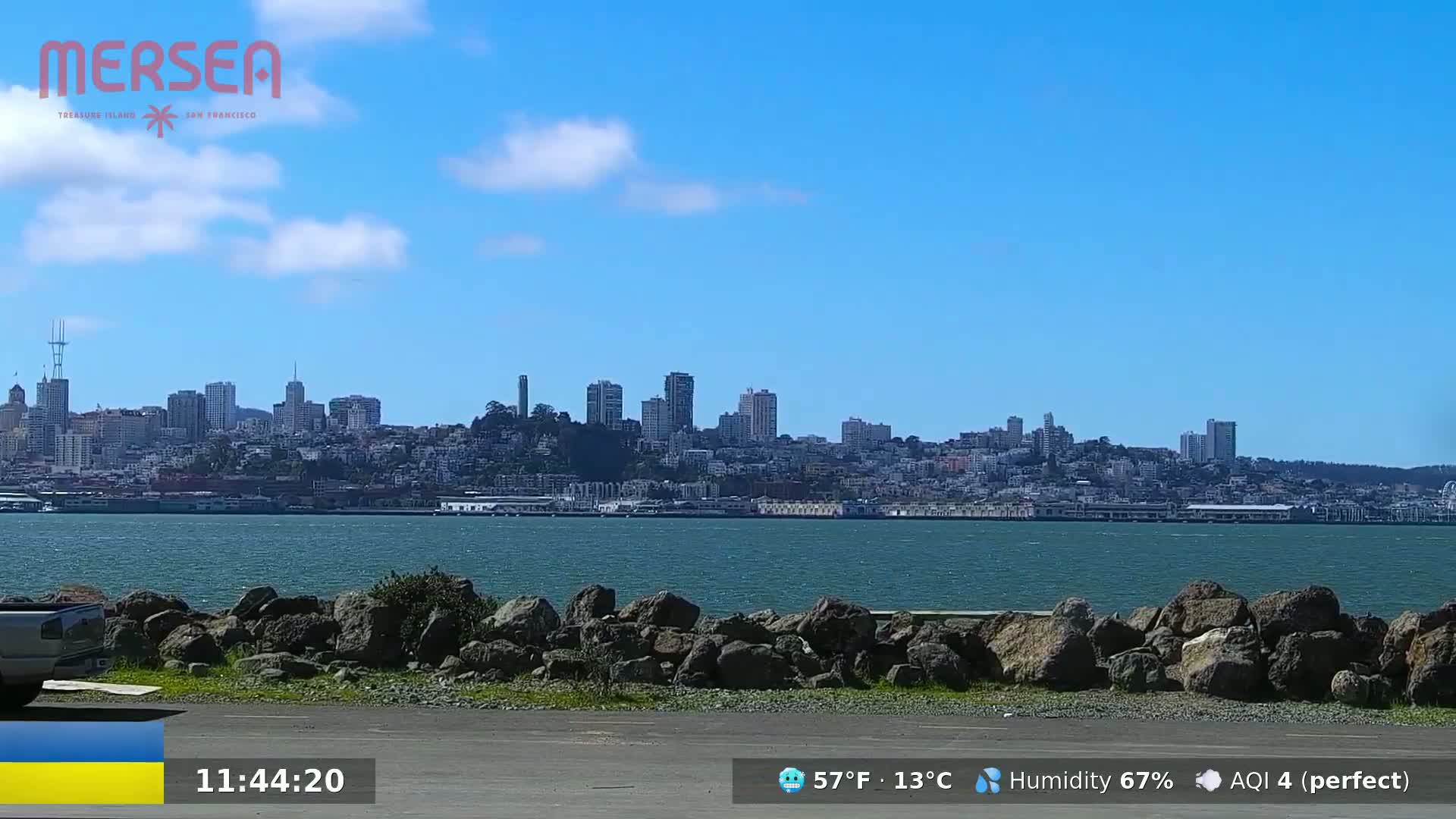 A city skyline is visible across a body of water on a sunny day with a few scattered clouds.