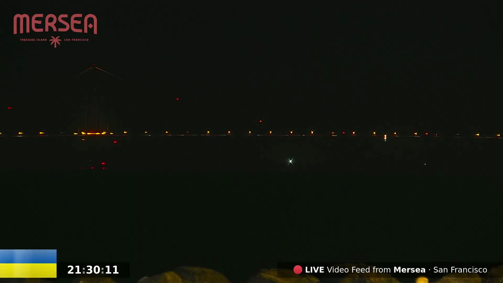 At night, a dark body of water reflects a line of lights, possibly from a bridge or pier, under a clear, dark sky.
