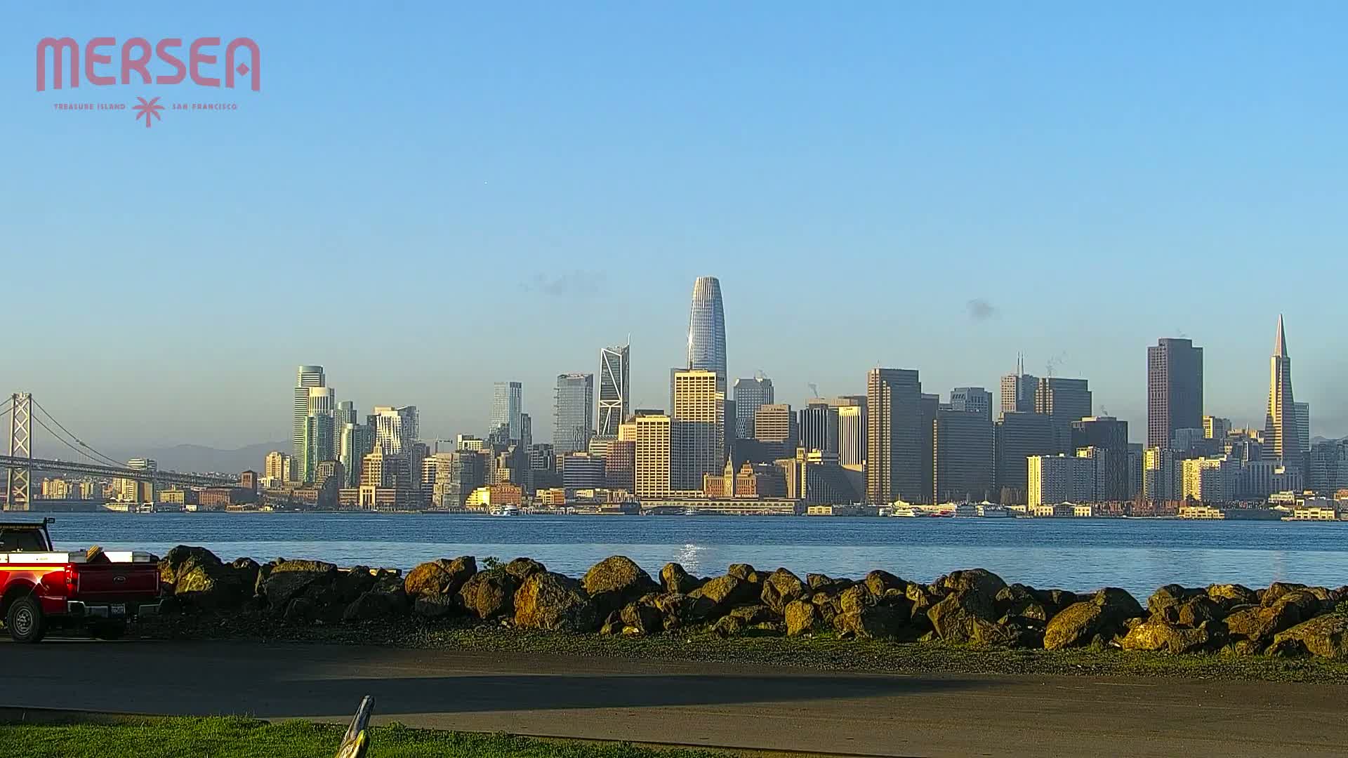 A red truck is parked near a rocky shoreline, with a clear view of the San Francisco skyline across a calm body of water under a bright, clear sky.