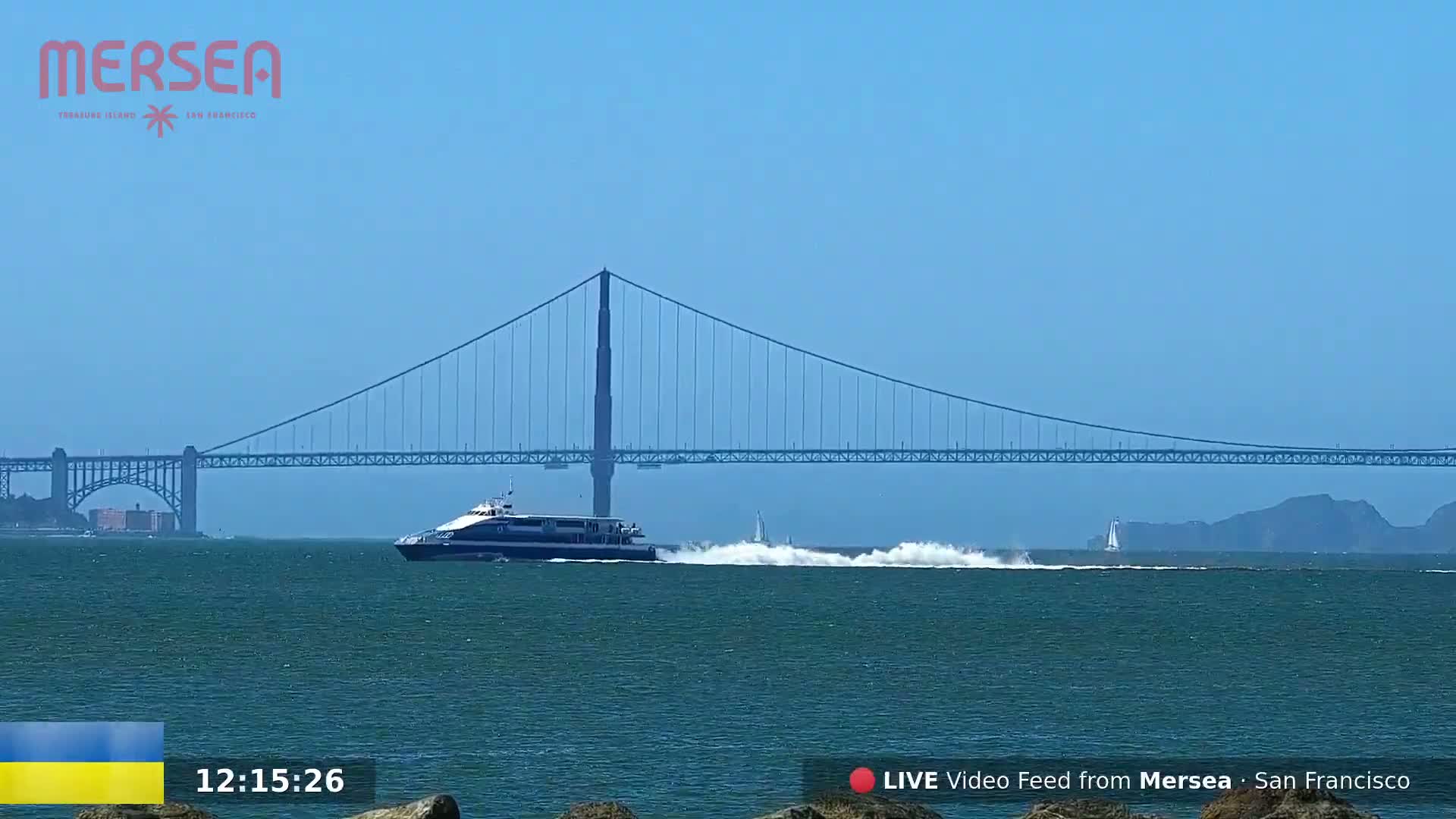 A ferry boat creates a wake as it travels across a body of water with a suspension bridge in the background under a clear blue sky.