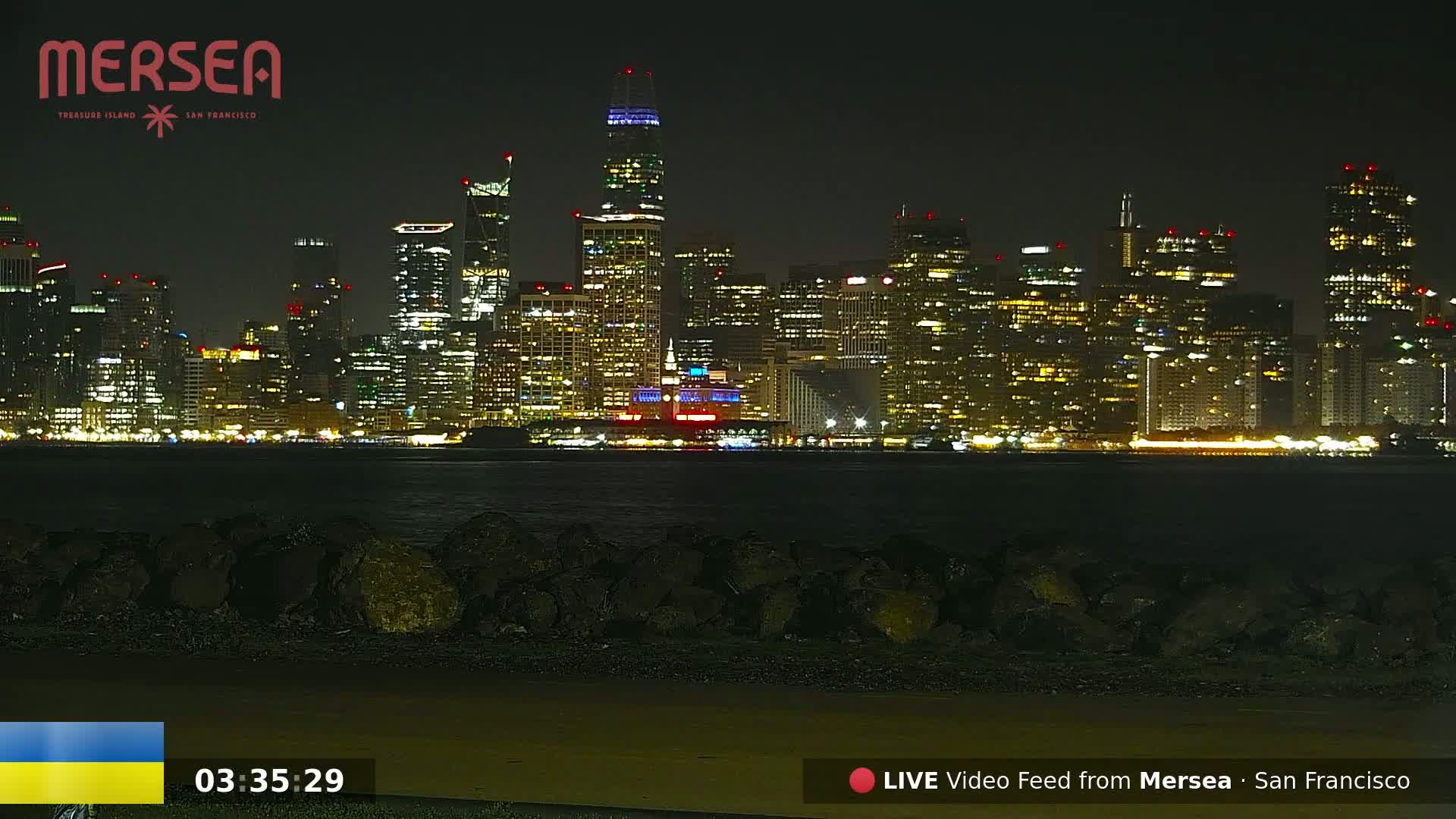 A nighttime view of a brightly lit city skyline across a dark body of water, with a rocky shoreline in the foreground.