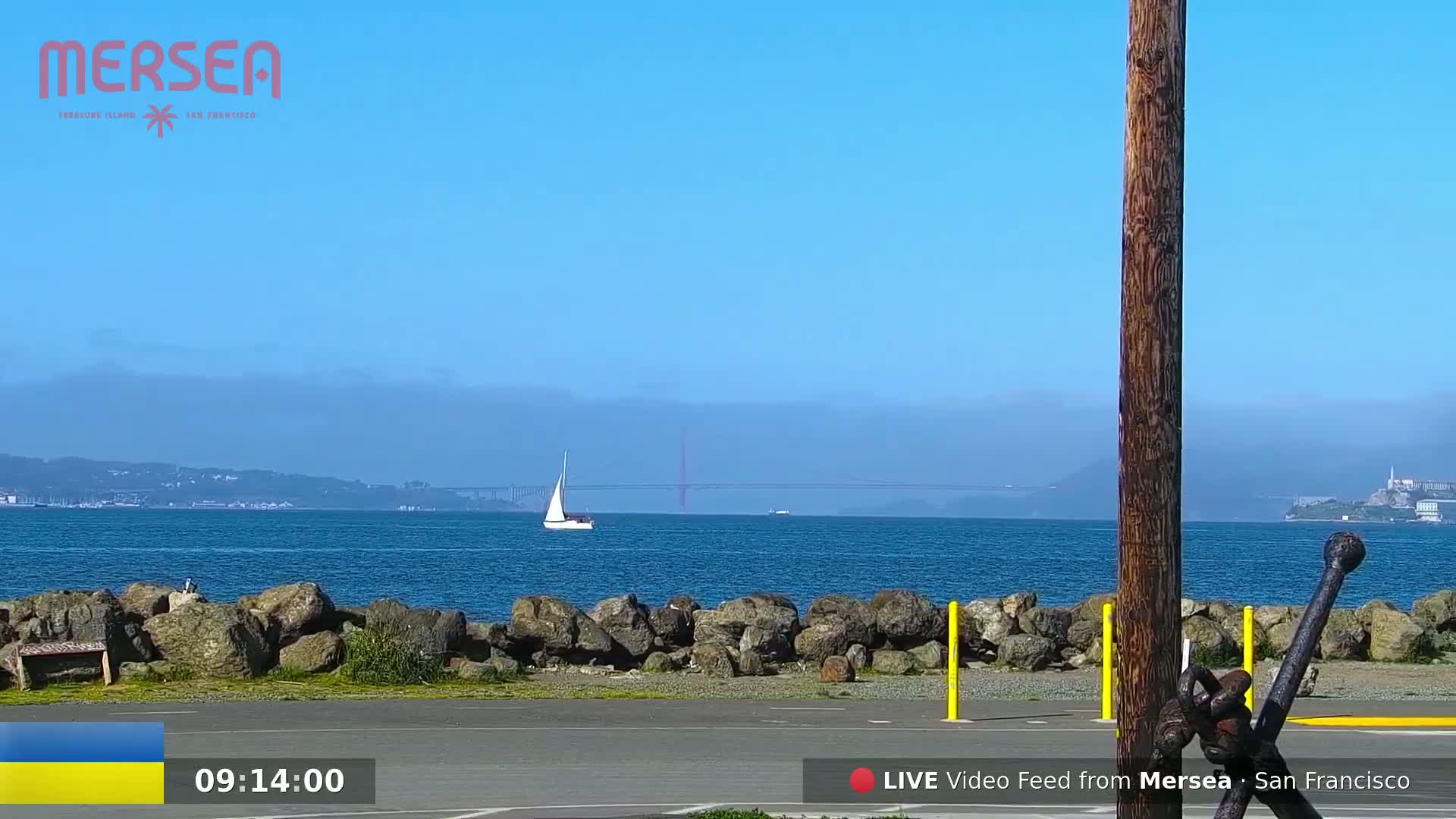 A sailboat is visible on a calm, blue body of water with a hazy background showing a bridge and distant land under a clear, bright blue sky.