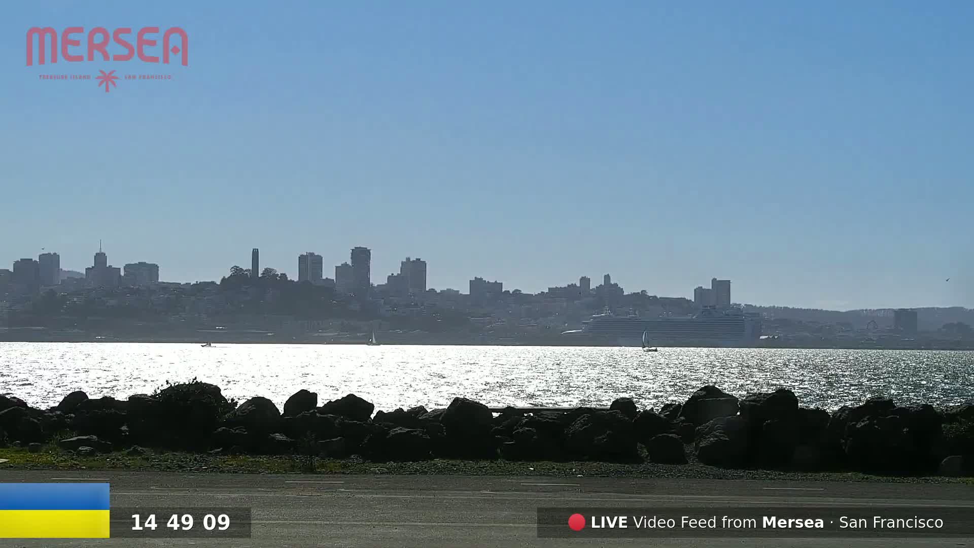 A city skyline is visible across a sunlit body of water, with dark rocks in the foreground under a clear blue sky.
