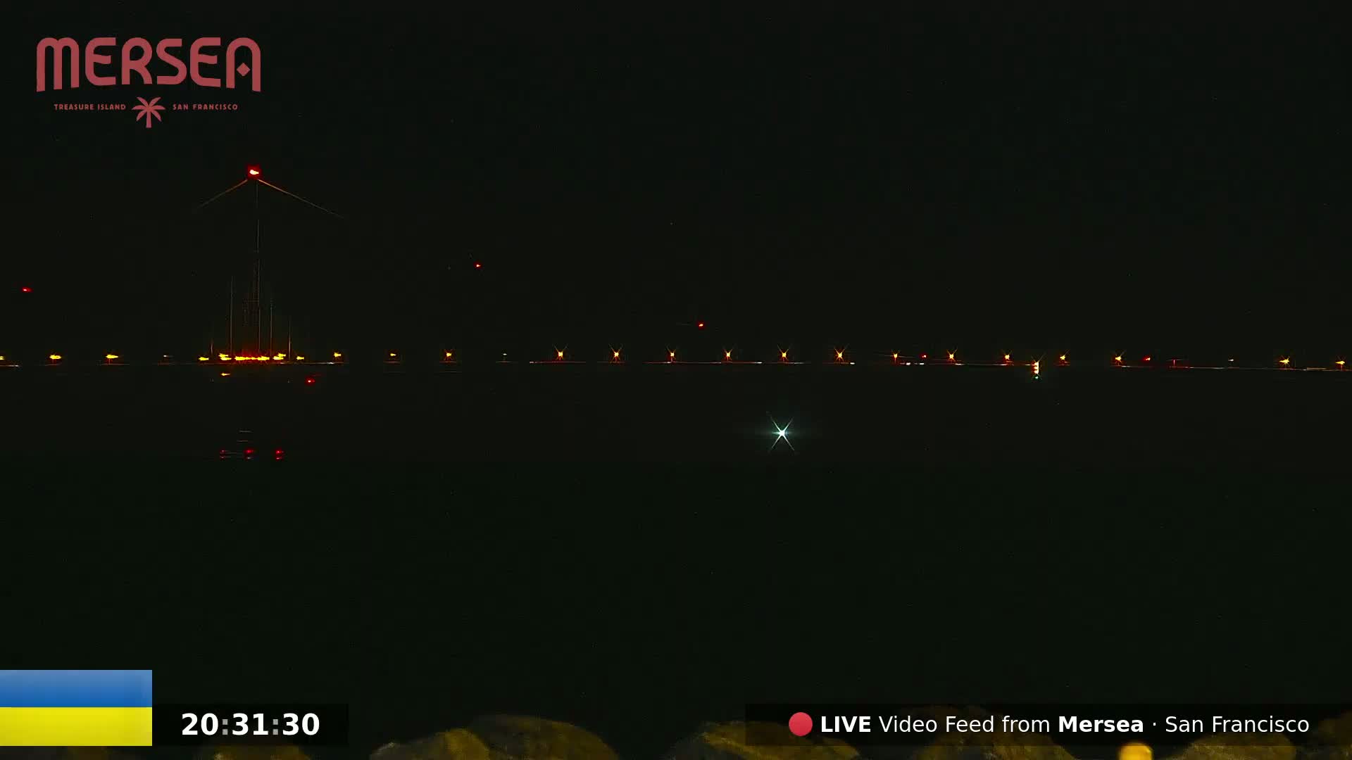 A dark nighttime scene shows a body of water reflecting a line of lights across the horizon, with a tall structure, possibly a tower, illuminated with red lights in the center.