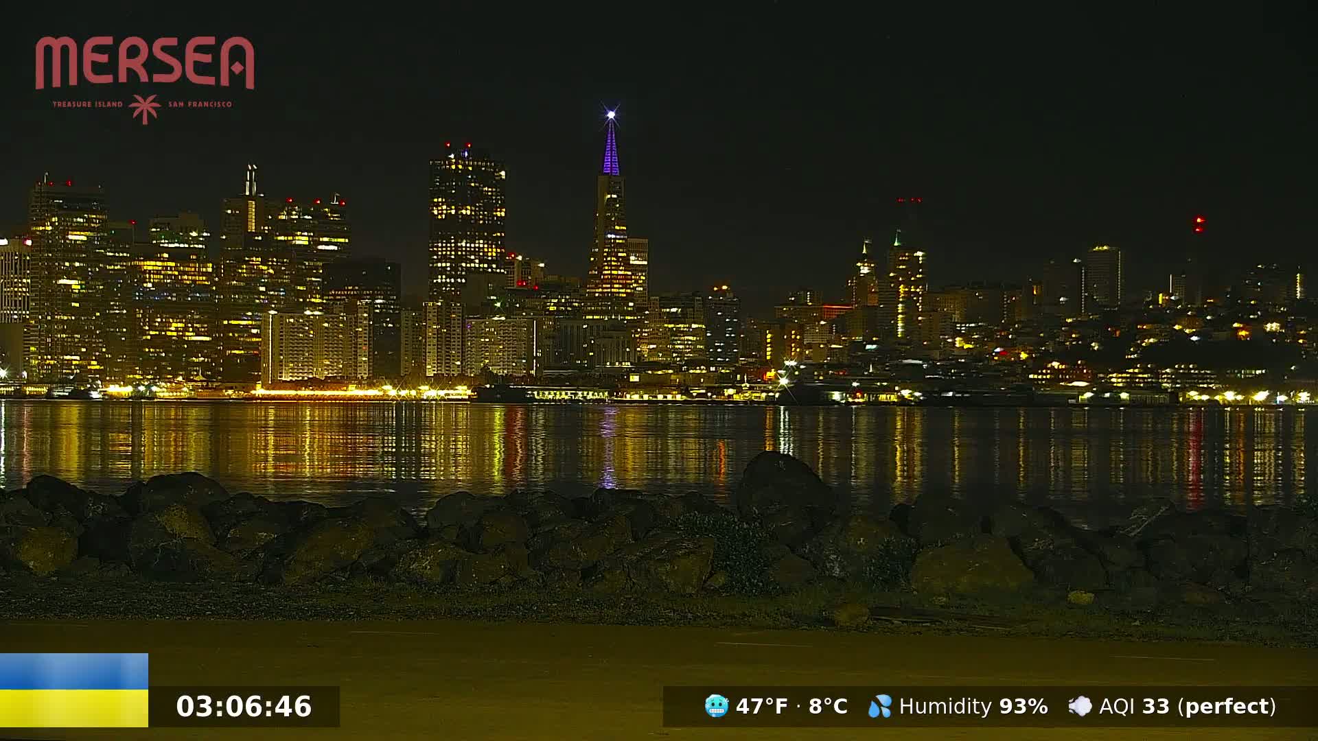 A nighttime view of a city skyline reflected in calm water, with a rocky shoreline in the foreground.