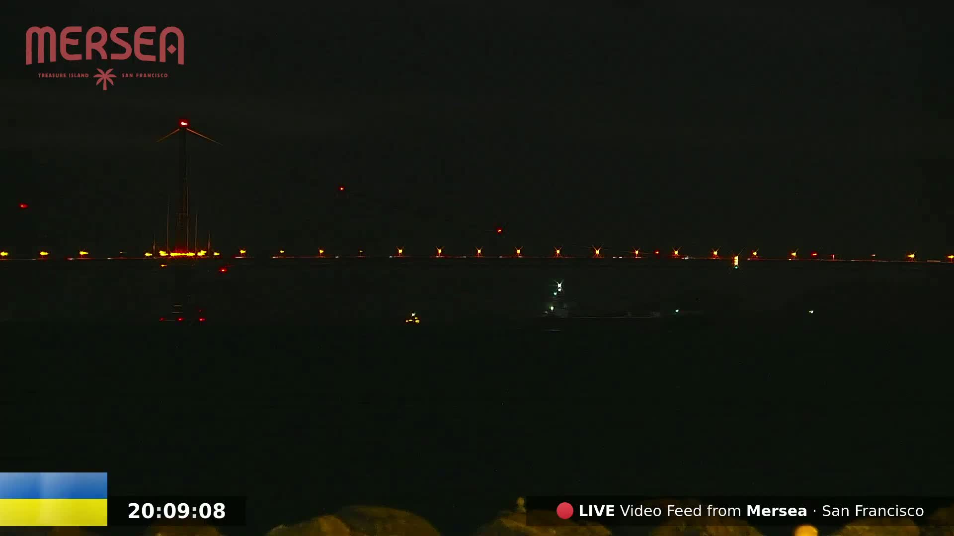A nighttime view of a body of water with a bridge in the distance illuminated by lights, a wind turbine, and a few small boats.