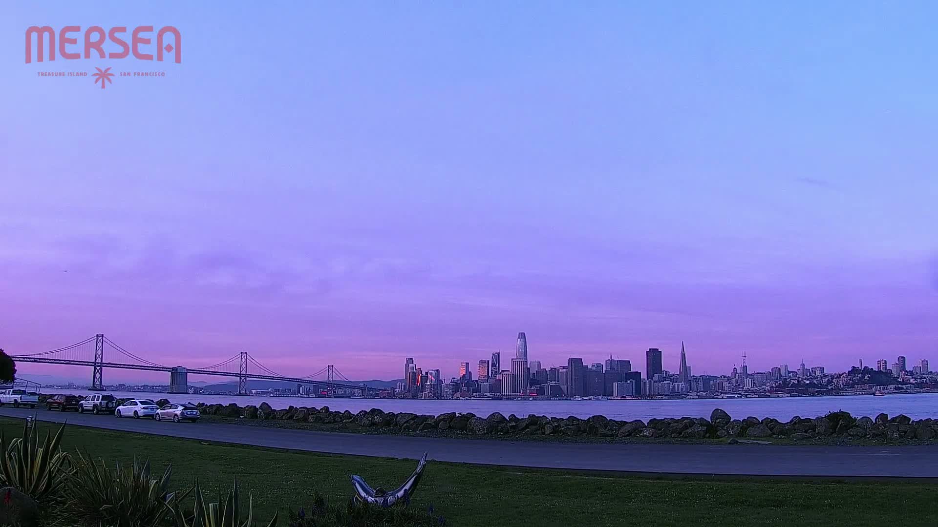 Under a purple-hued twilight sky, a cityscape is seen across a body of water from a grassy area with parked cars and a sculpture.