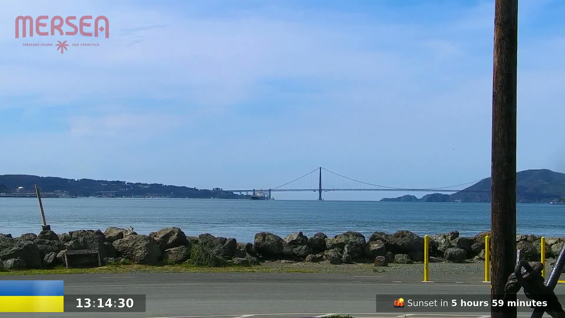 A suspension bridge is visible in the distance across a calm body of water under a clear, bright sky, with a rocky shoreline in the foreground.
