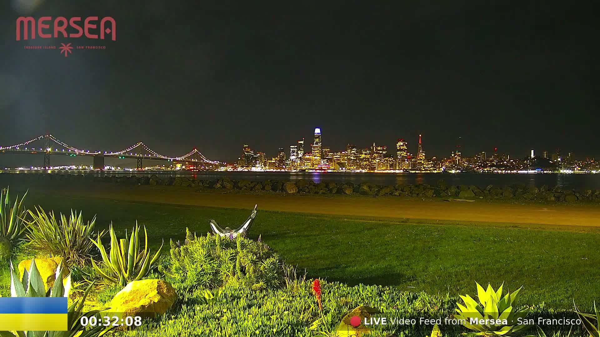 A nighttime view of a city skyline across a body of water, with a bridge in the foreground and lush green landscaping in the immediate foreground.