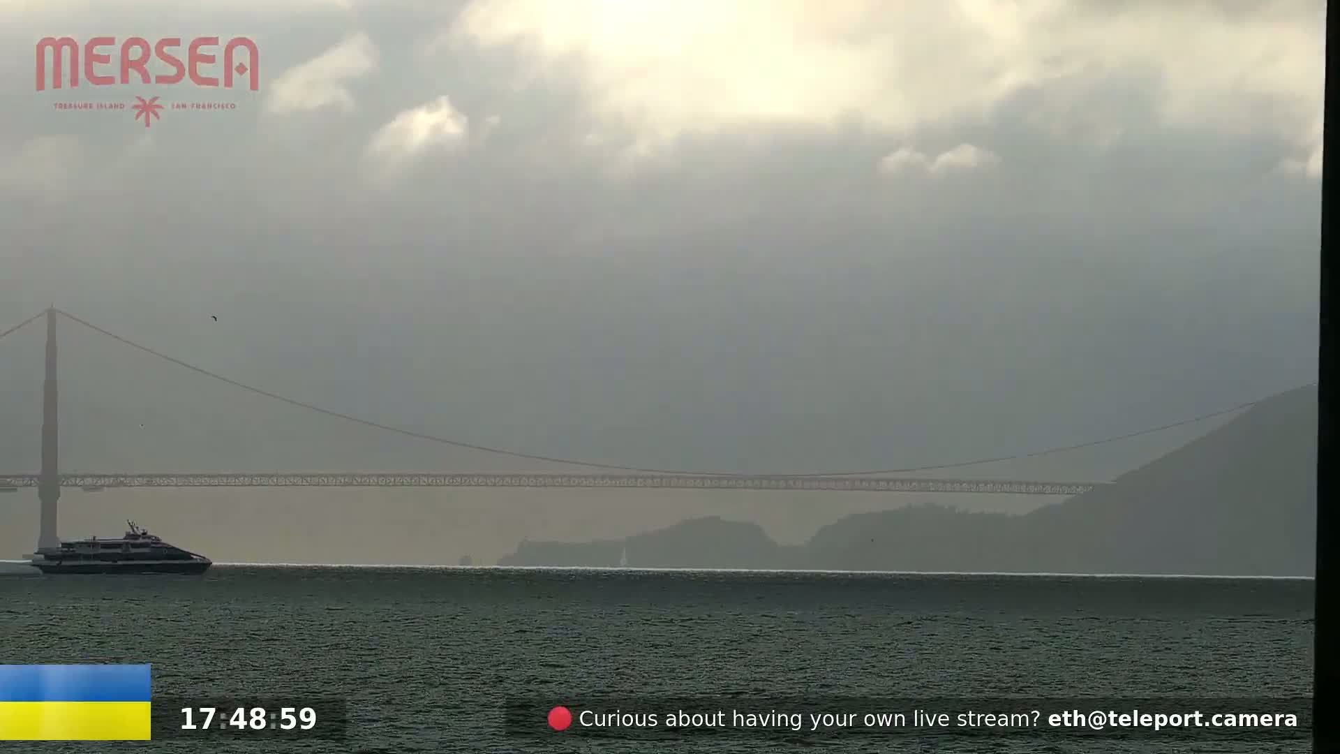 A ferry travels across a body of water under an overcast sky, with a suspension bridge in the background.