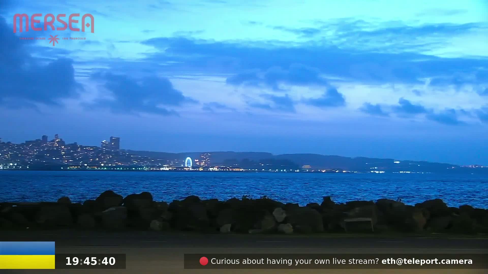 A nighttime view of a city skyline across a dark blue body of water, under a mostly clear, twilight sky.