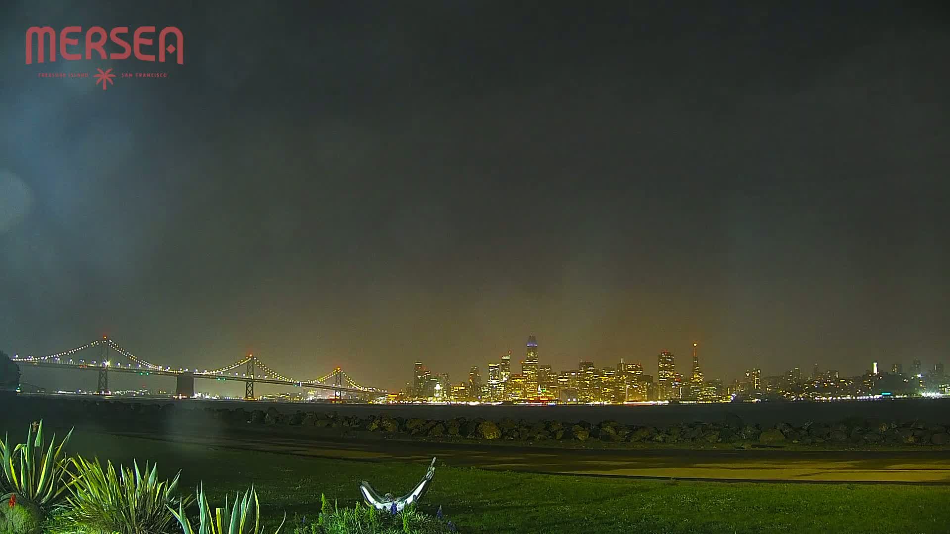 A nighttime view of a cityscape across a body of water, with a suspension bridge in the foreground under a hazy sky.